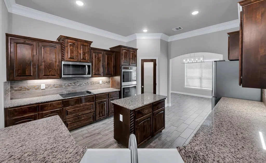 Kitchen featuring wood cabinetry, light-colored granite countertops, stainless steel appliances, a tile backsplash, and a central island