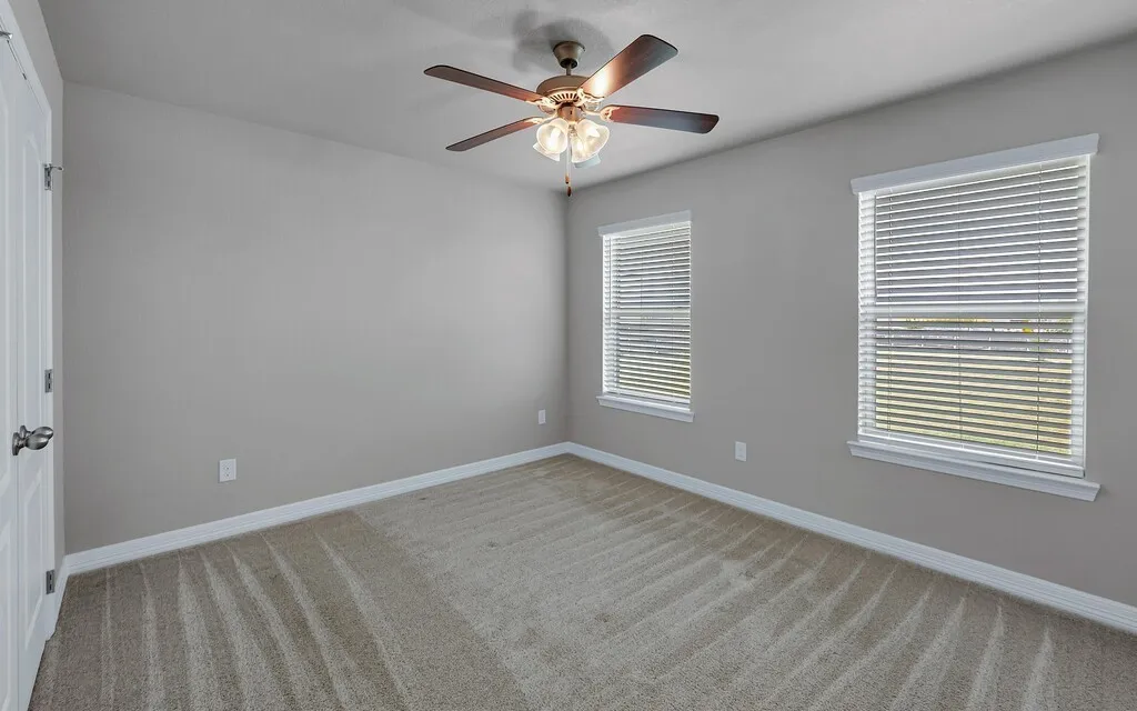 Neutral-toned walls, two windows with blinds, ceiling fan with integrated lighting, and carpeted flooring