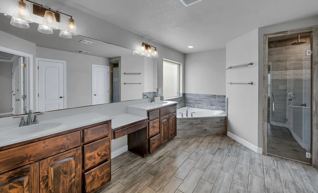 Dual vanities with integrated sinks, a built-in vanity station, a corner soaking tub, a separate glass-enclosed shower, and wood-finish tile flooring