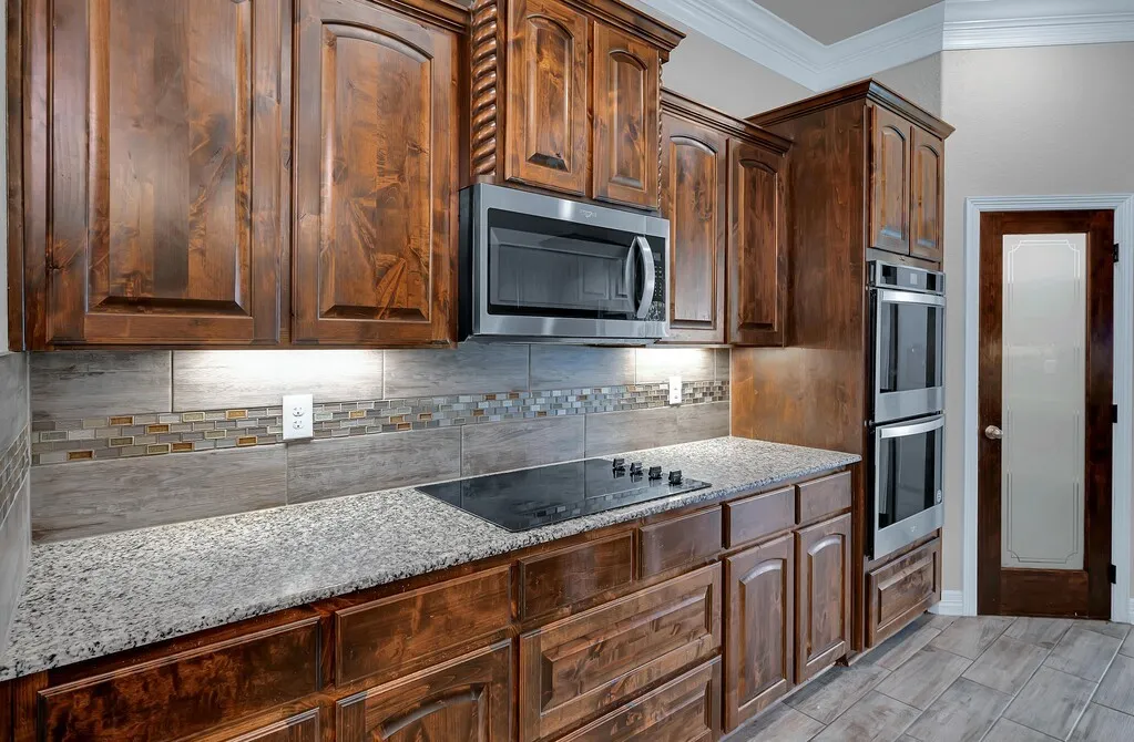Warm wood cabinetry with crown molding, stainless steel microwave, integrated cooktop, stone countertops, and tile backsplash with decorative mosaic accent