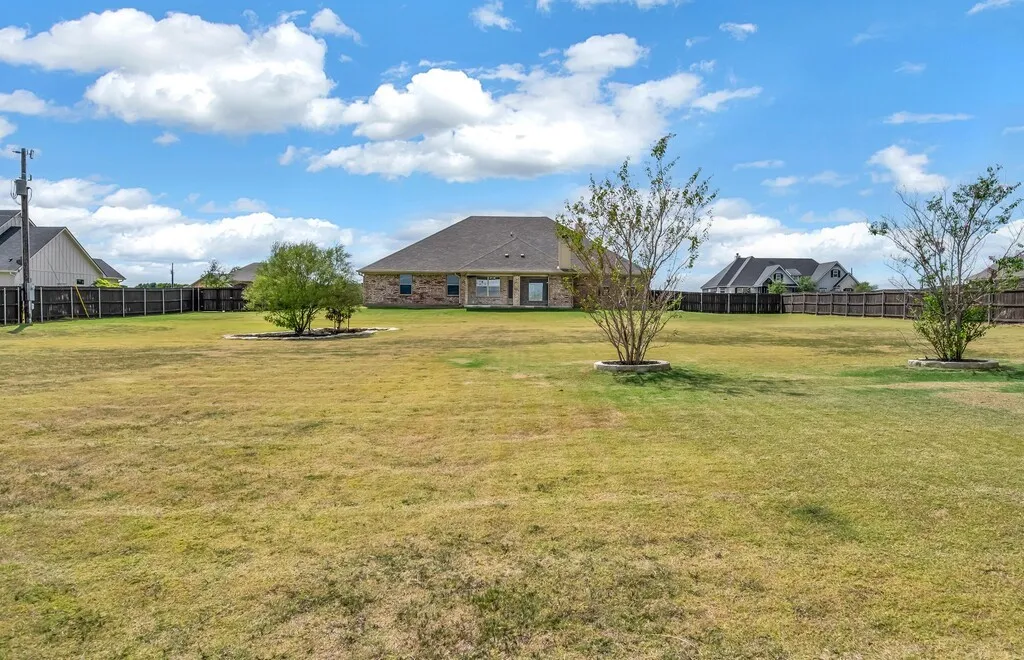 Expansive backyard featuring a privacy fence, mature trees, and a brick exterior dwelling with a shingle roof