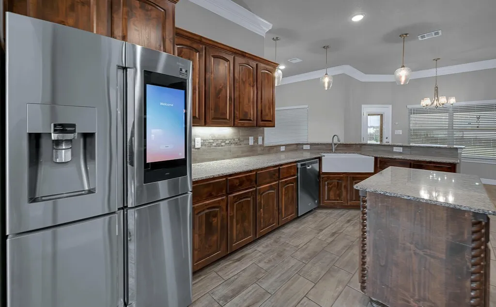 Kitchen featuring wood-finish cabinetry, stone countertops, a large farmhouse sink, a stainless steel refrigerator, and wood-look tile flooring