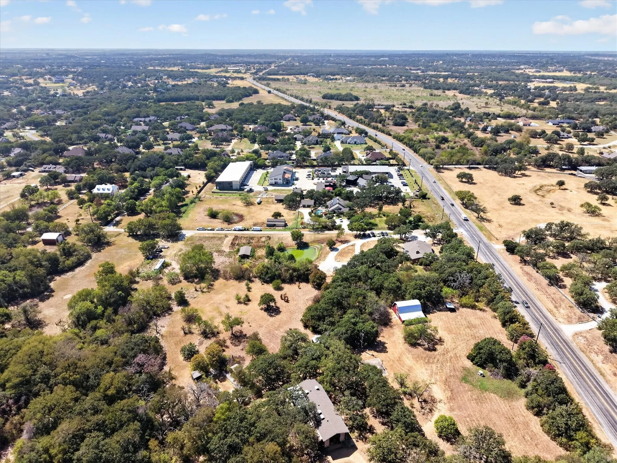 Wide aerial perspective highlighting the property, mature trees, open spaces, and nearby roadway.