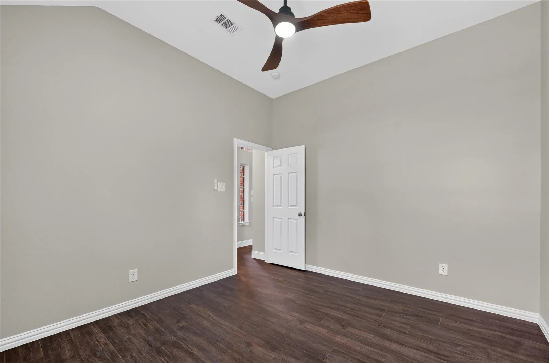 Spacious room featuring wood-finish flooring, light gray walls, a white paneled door, and an angled ceiling with a ceiling fan