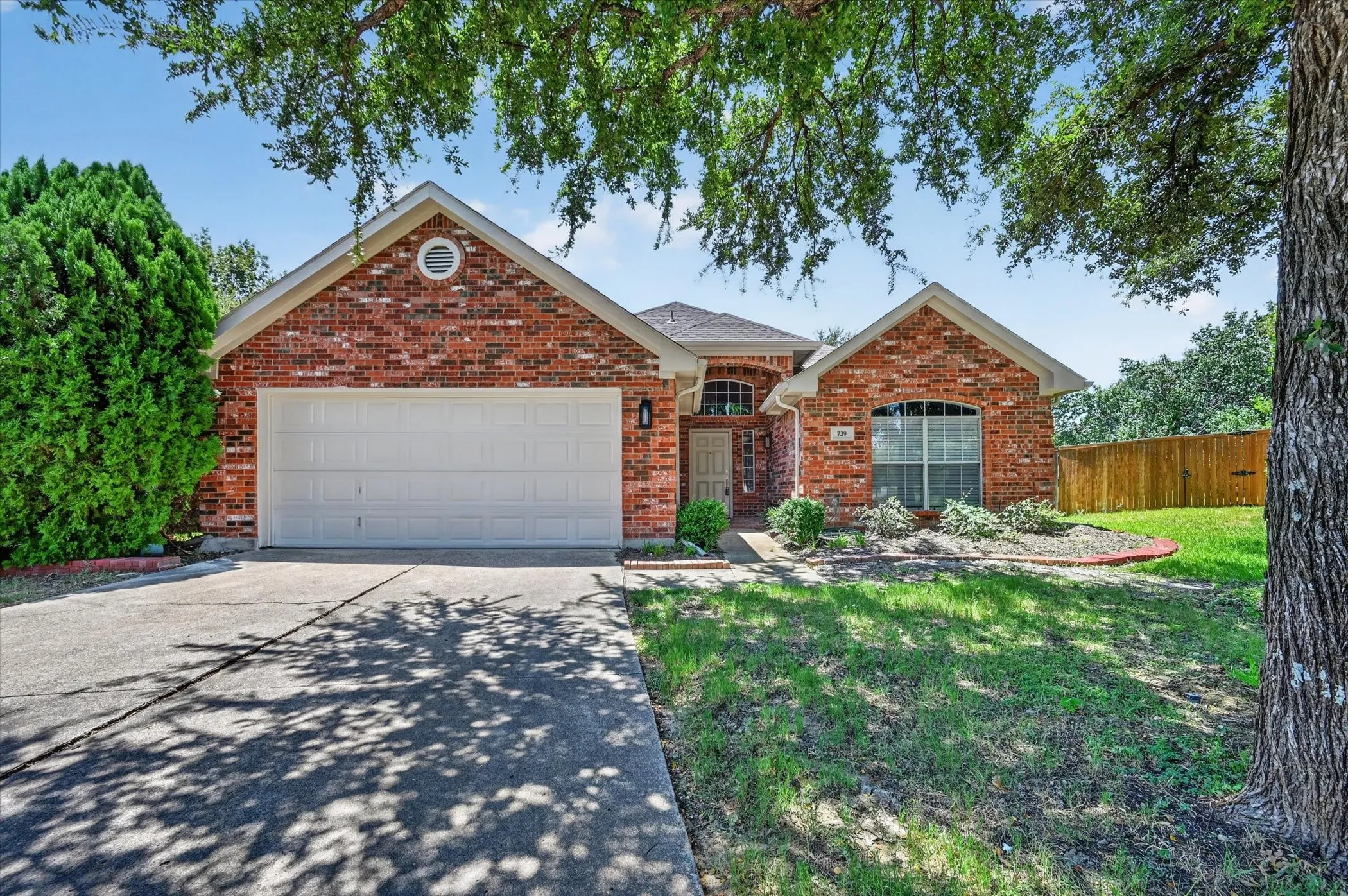 Brick exterior, attached garage, concrete driveway, arched front window, and manicured front landscaping