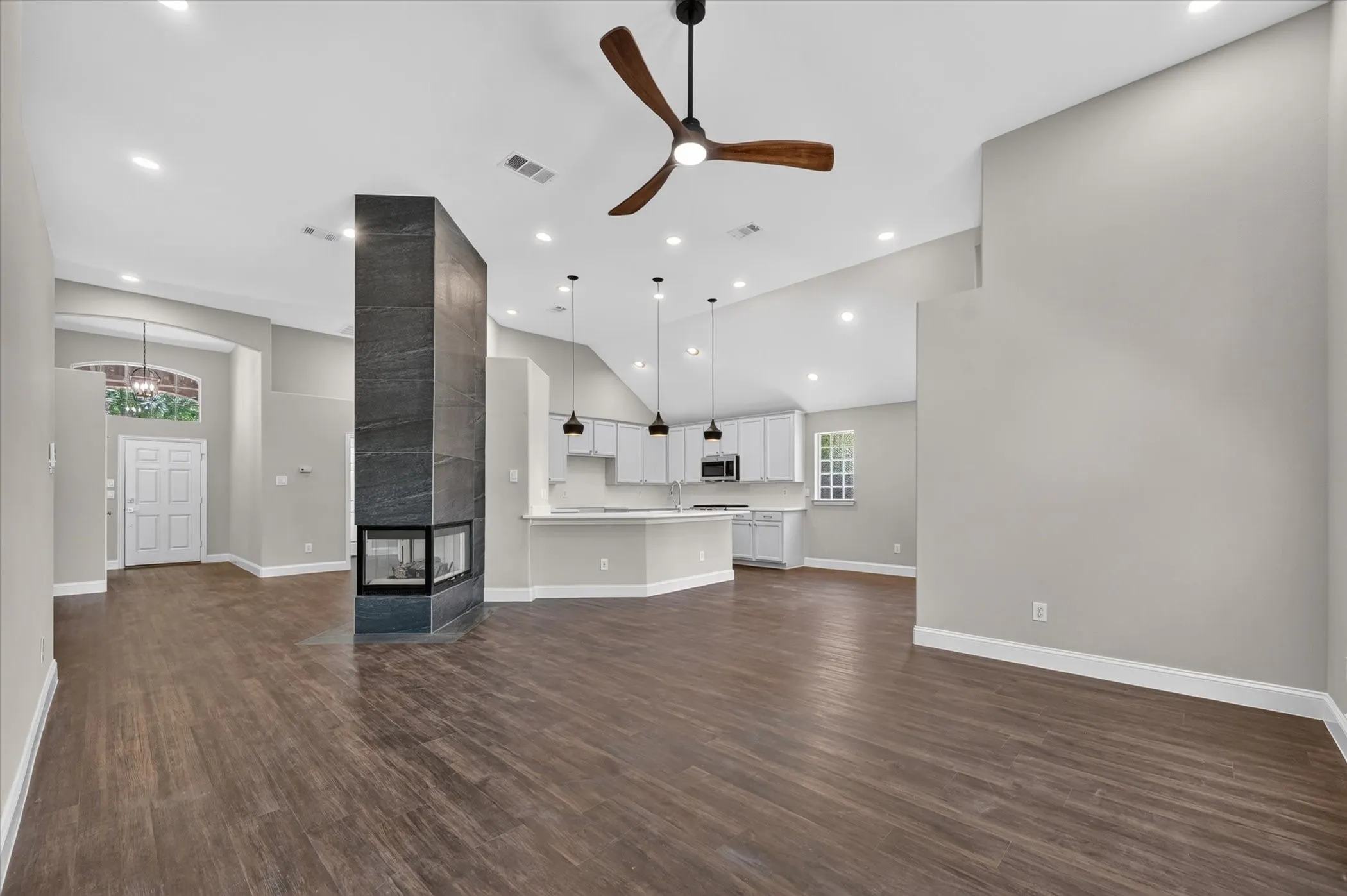 Wood-finish flooring throughout, a central fireplace with a dark stone-clad surround, a kitchen with white cabinetry, and recessed lighting