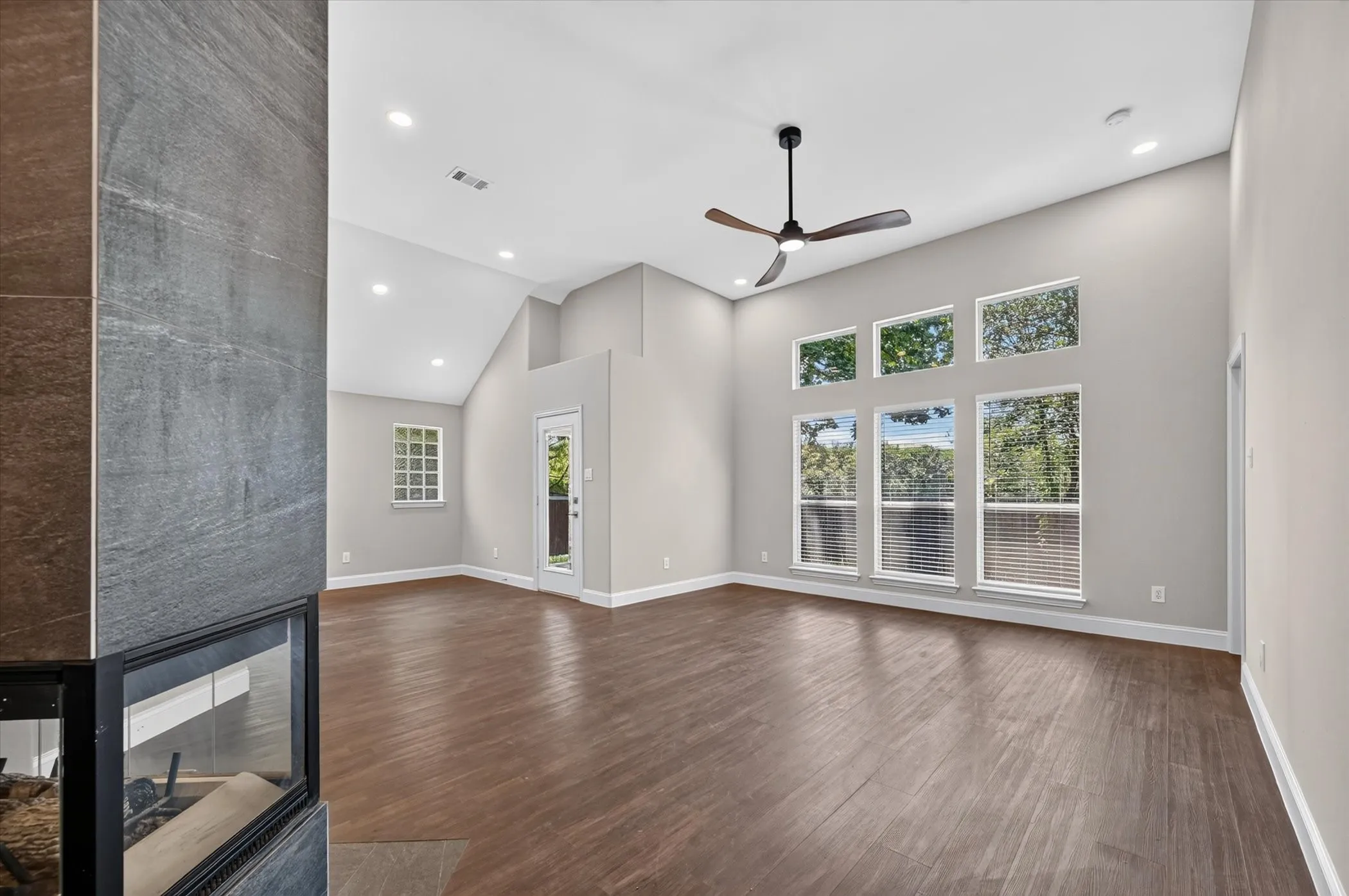 Living area featuring wood-finish flooring, a contemporary fireplace with a dark grey stone surround, a high vaulted ceiling, recessed lighting, and a modern ceiling fan
