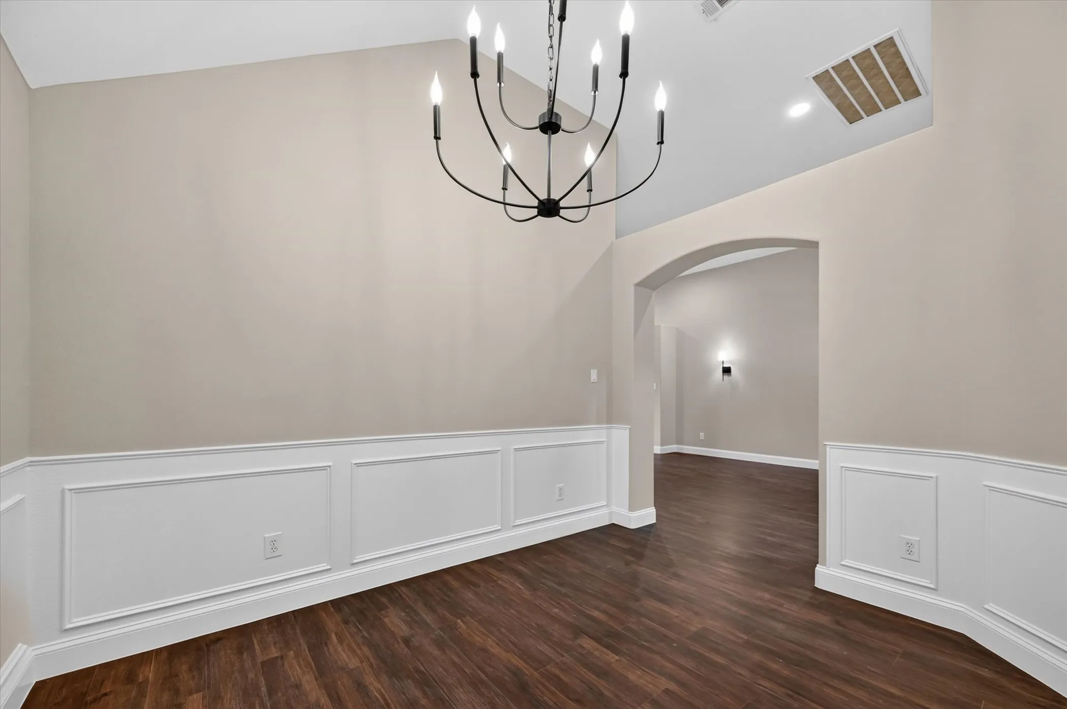 Wood-finish flooring, wainscoting, vaulted ceiling, arched doorway, and a black chandelier