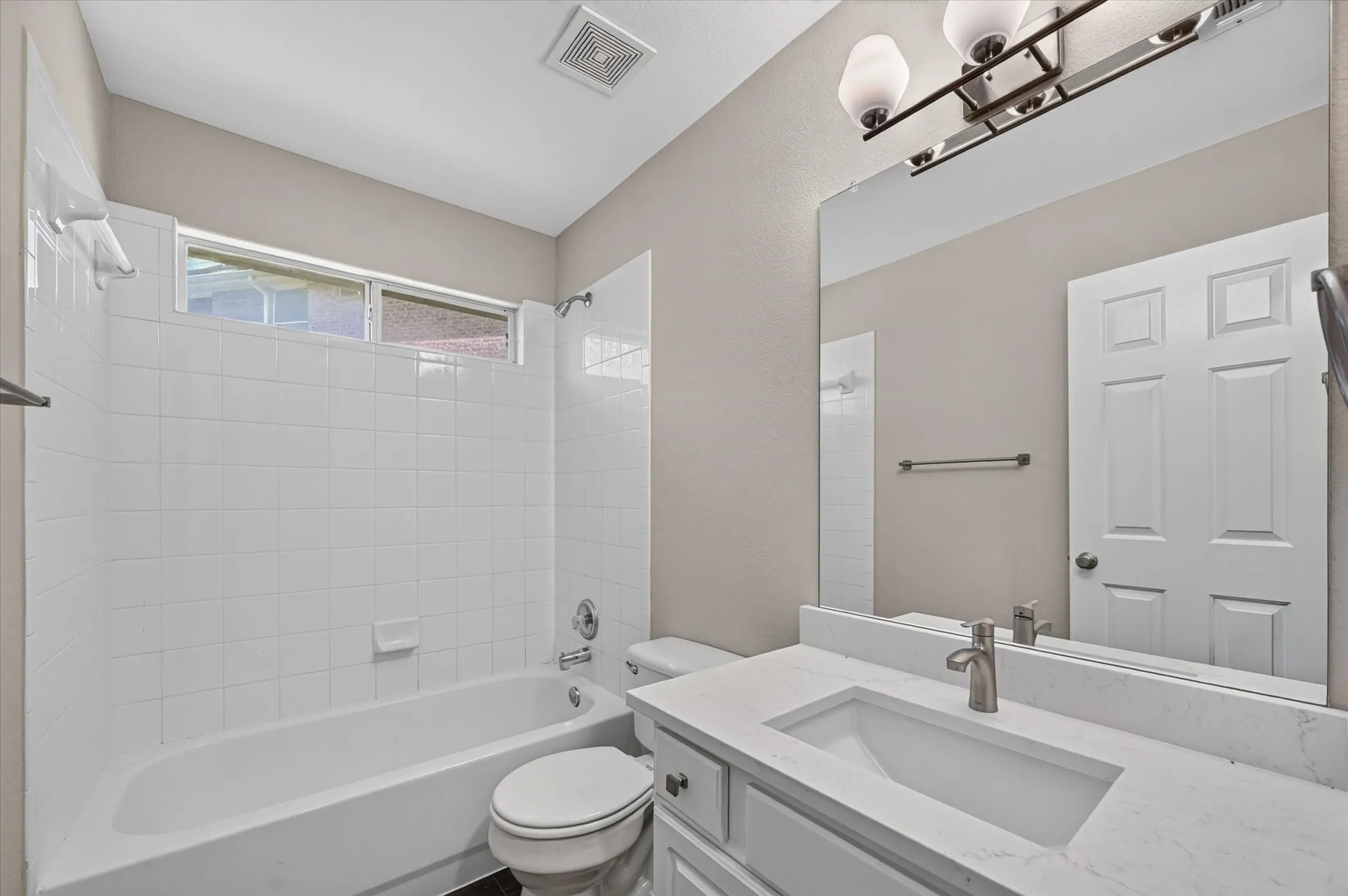 White tiled tub/shower combination, above-tub window, vanity with integrated sink, rectangular mirror, and a three-light fixture