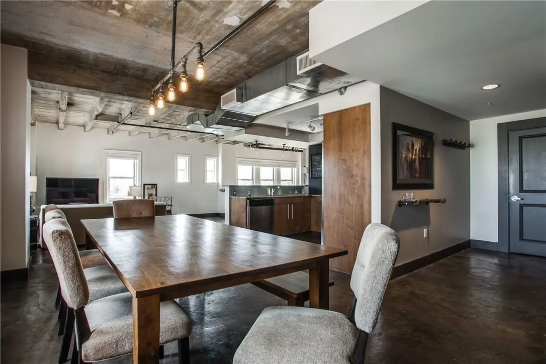 Exposed concrete ceiling and structural beams, track lighting with Edison bulbs, wood-finish flooring, and multiple windows