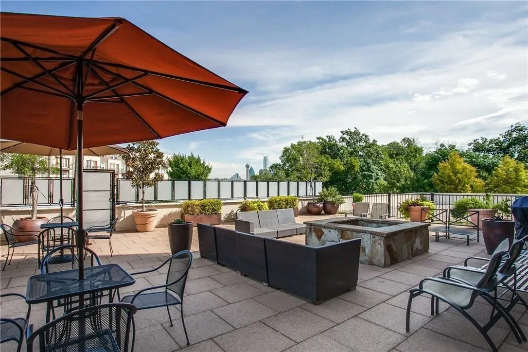 Expansive paver patio featuring a built-in stone fire pit, metal railing, and various planted trees and shrubs