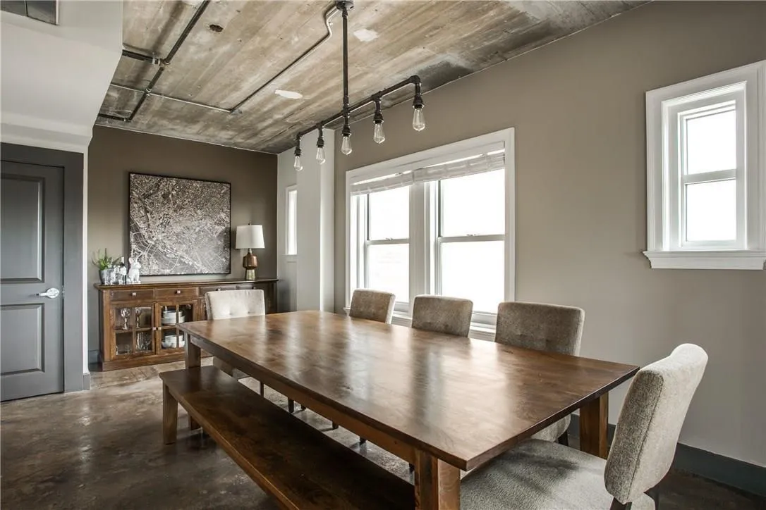 Exposed concrete ceiling with integrated piping, multiple windows with white trim, dark wood-finish flooring, and a dark-paneled door