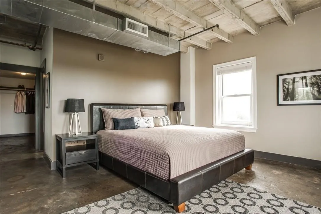 Exposed concrete ceiling with structural beams, industrial-style ductwork, single-pane window with white trim, concrete flooring, and an open closet with built-in hanging rods