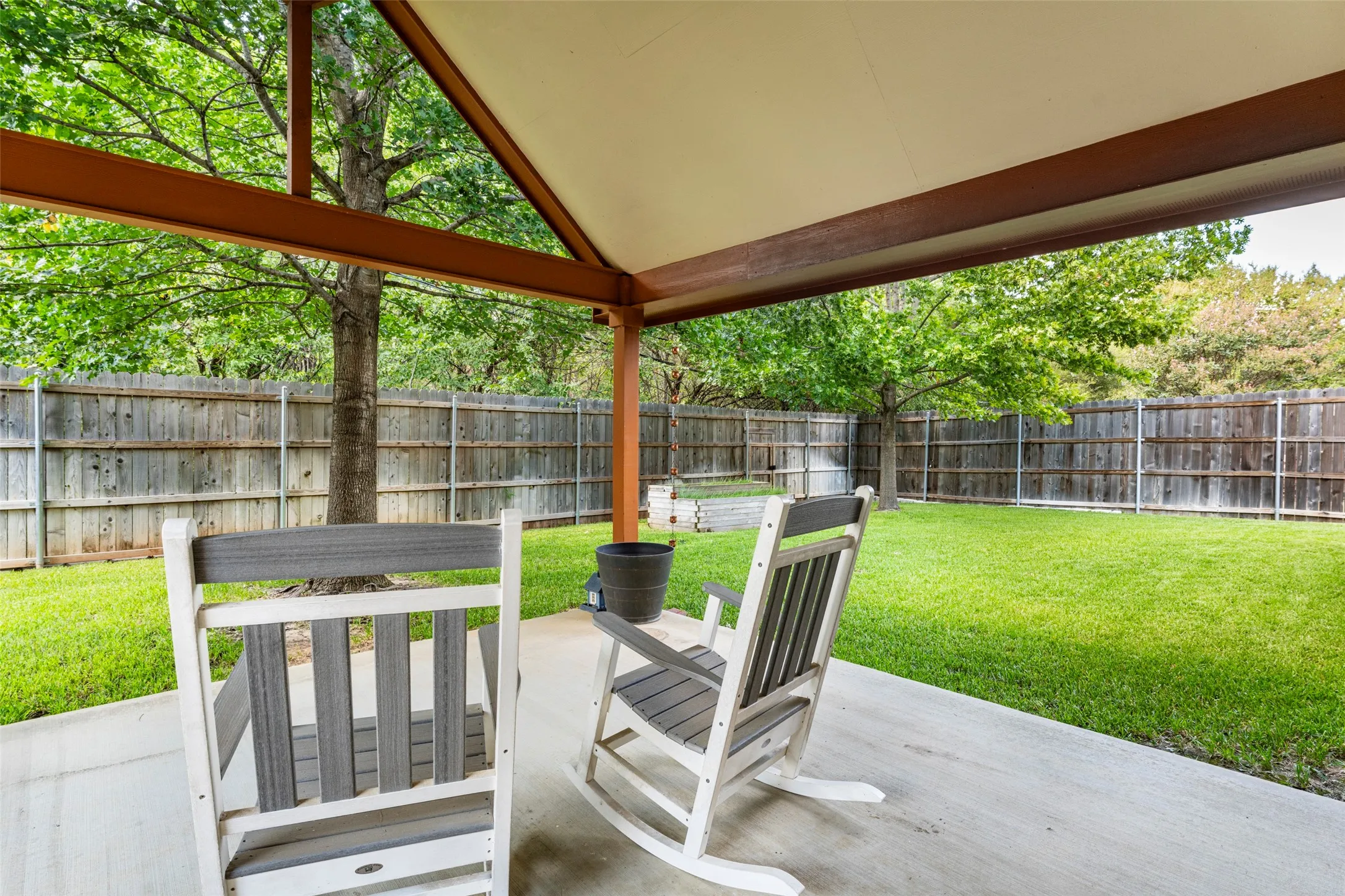 15x15 Covered patio with concrete flooring, supported by wood beams, overlooking a fenced backyard with a grass lawn and mature trees