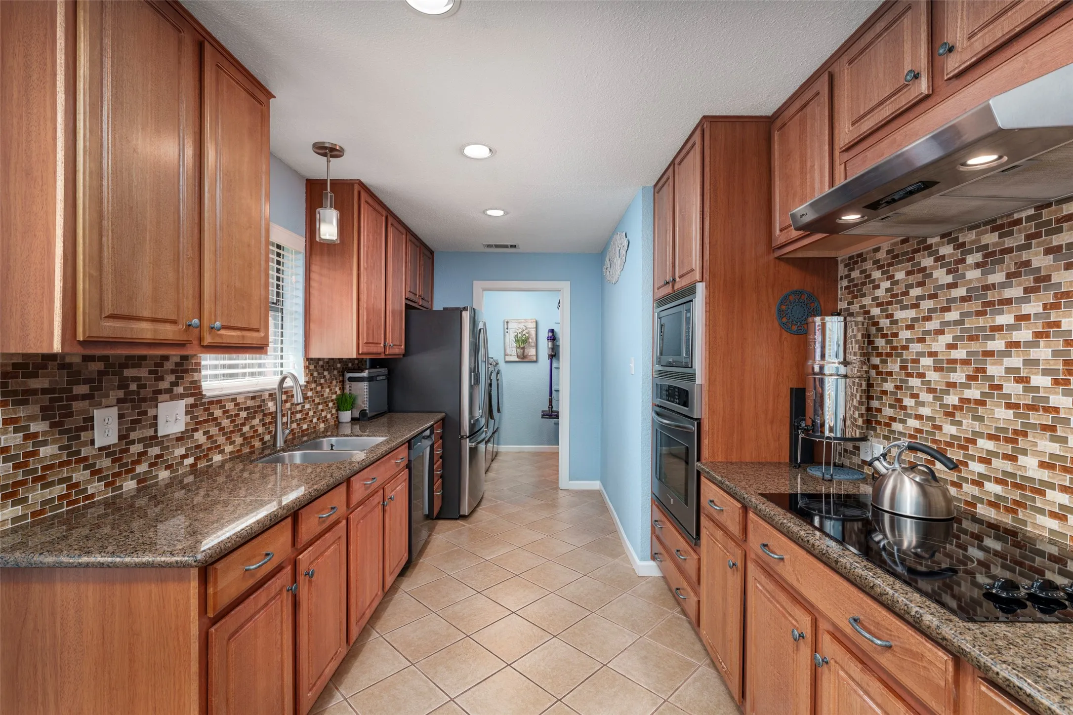 Galley kitchen featuring wood cabinetry, stone countertops, mosaic tile backsplash, stainless steel appliances, and recessed lighting & Motion Sensor faucet