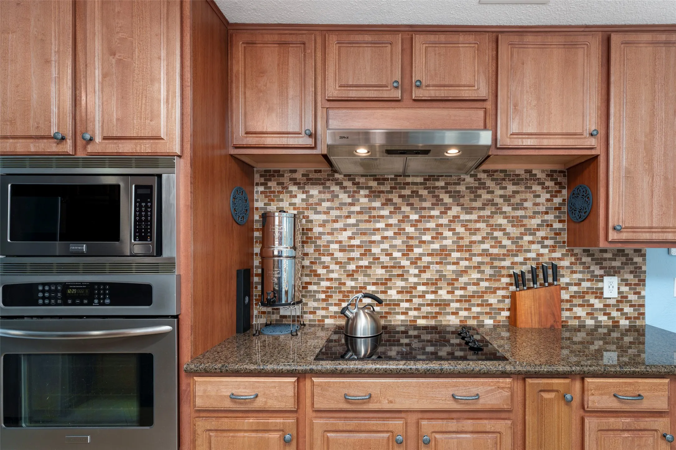 Built-in stainless steel microwave and oven, wood cabinetry, mosaic tile backsplash, and dark speckled countertop. Motion Sensor faucet