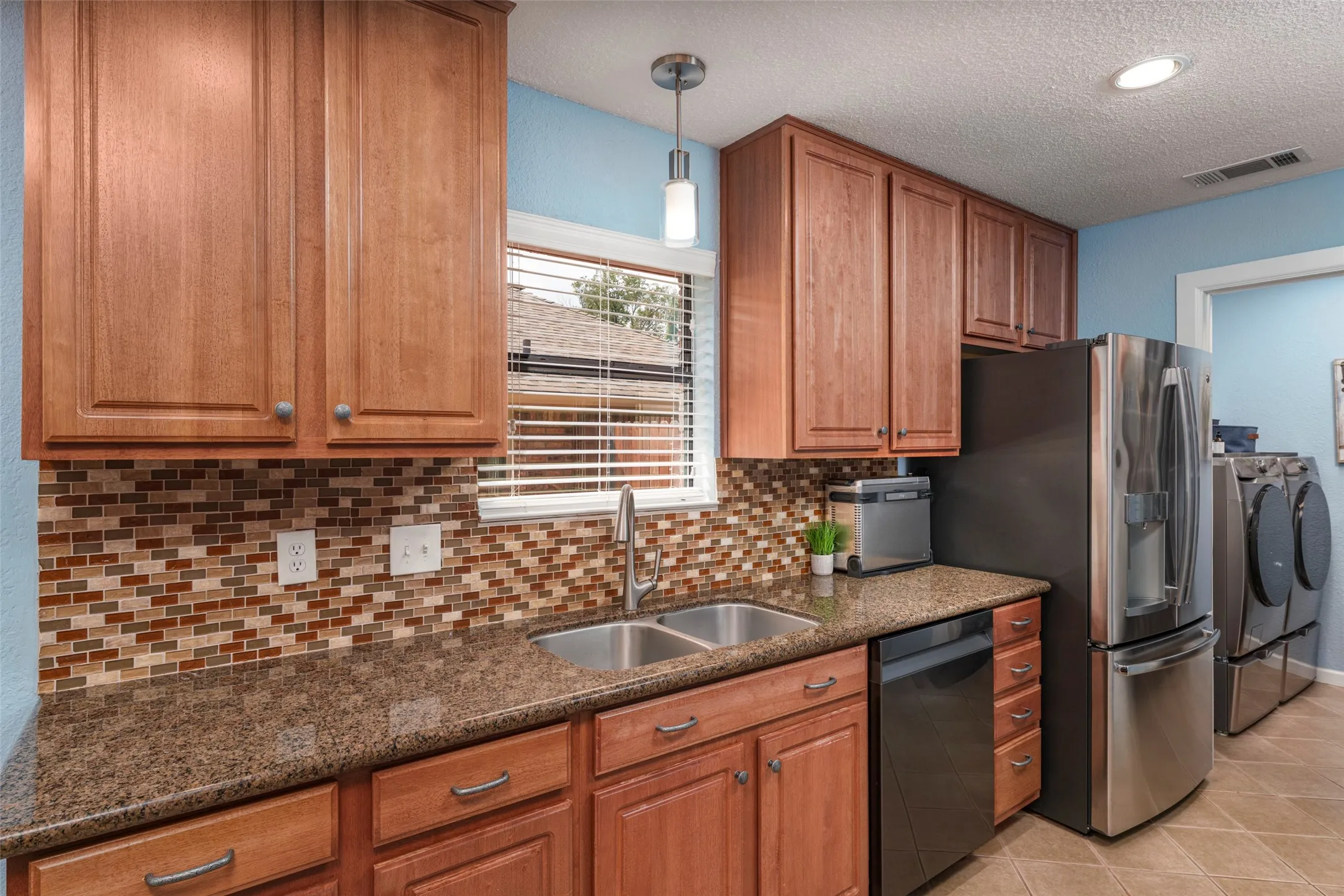 Wood-finish cabinetry, mosaic tile backsplash, durable countertop, double basin sink, and integrated stainless steel appliances. Motion Sensor faucet