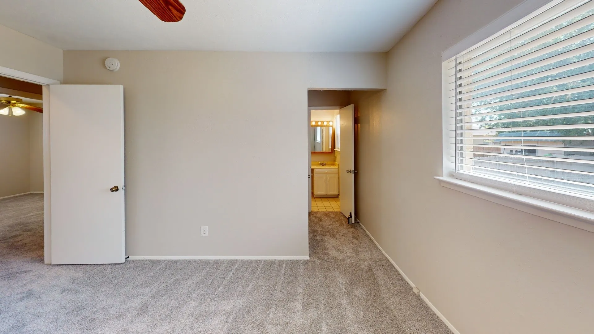 Plush carpet flooring, neutral paint tones, window with horizontal blinds, and a wood-finish ceiling fan