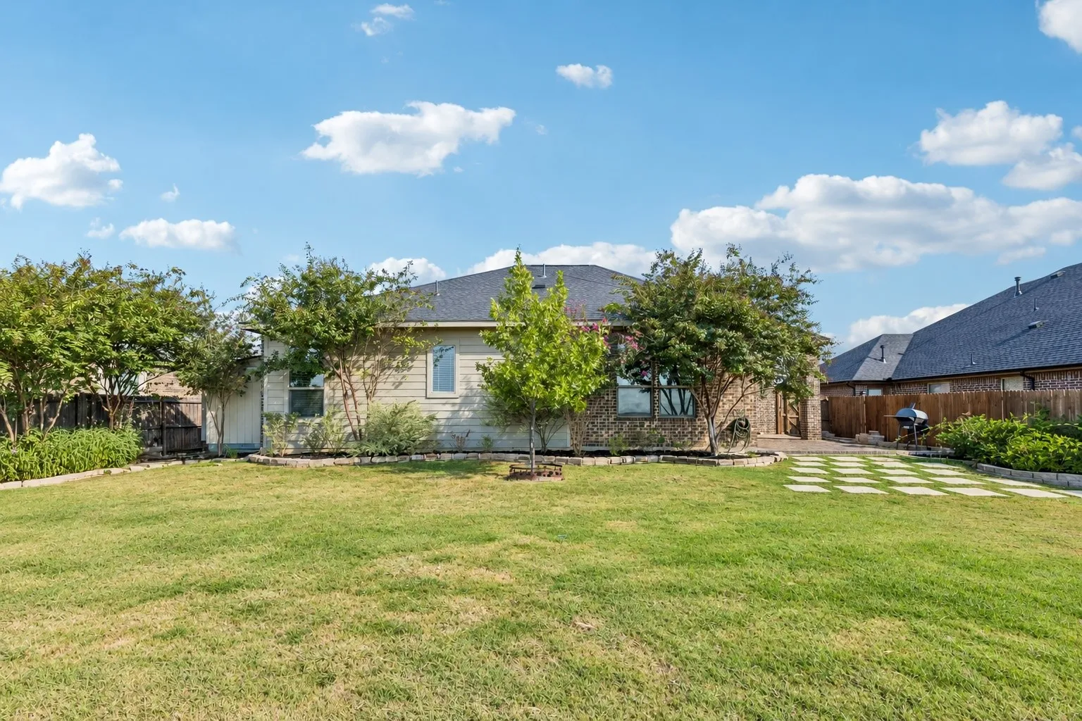 Expansive backyard featuring a manicured lawn, landscaping beds with mature trees, and a stone paver patio