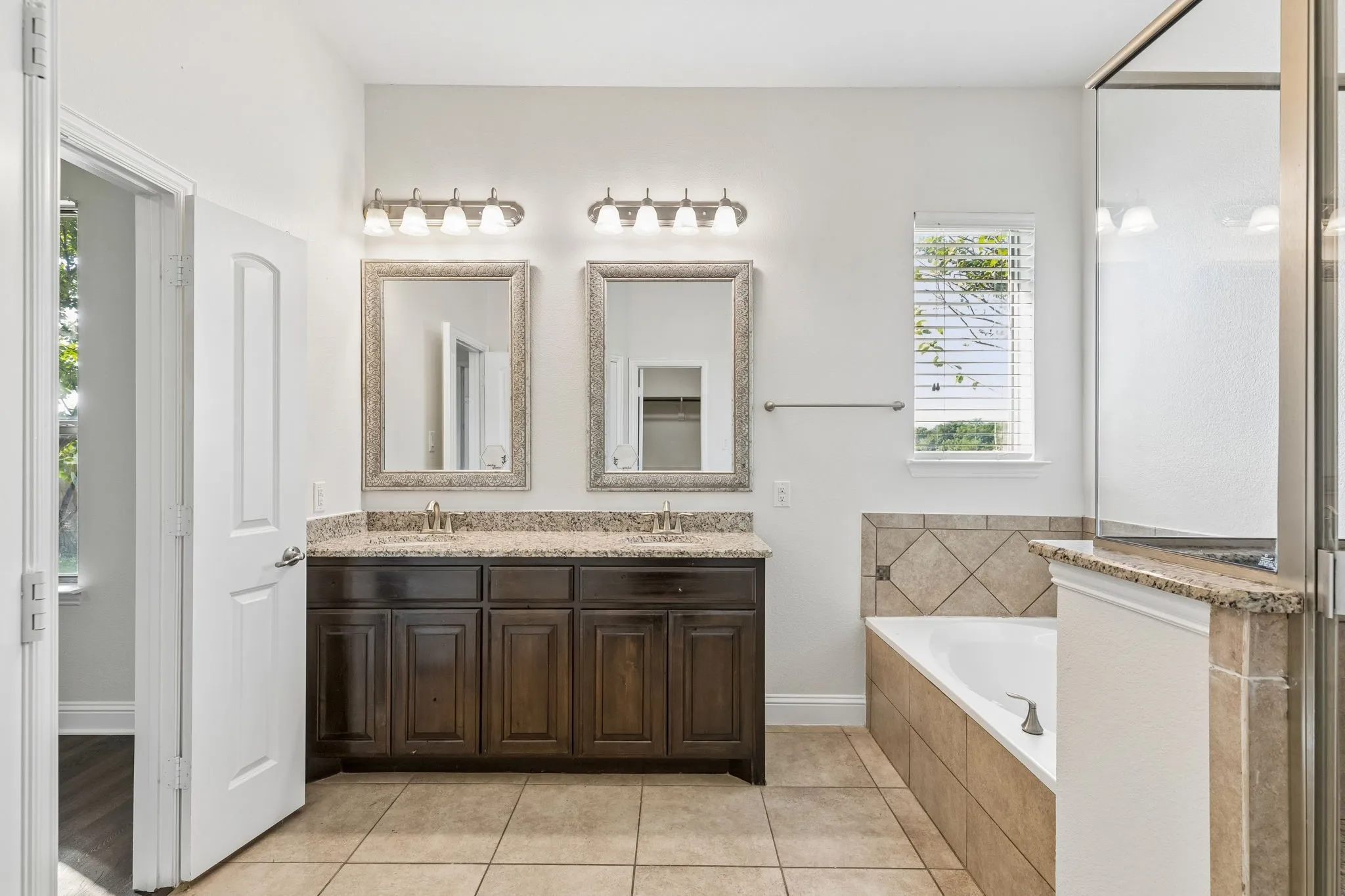 Dual vanity with dark wood-finish cabinetry and stone-finish countertops, framed mirrors, tiled flooring, and a tiled tub surround