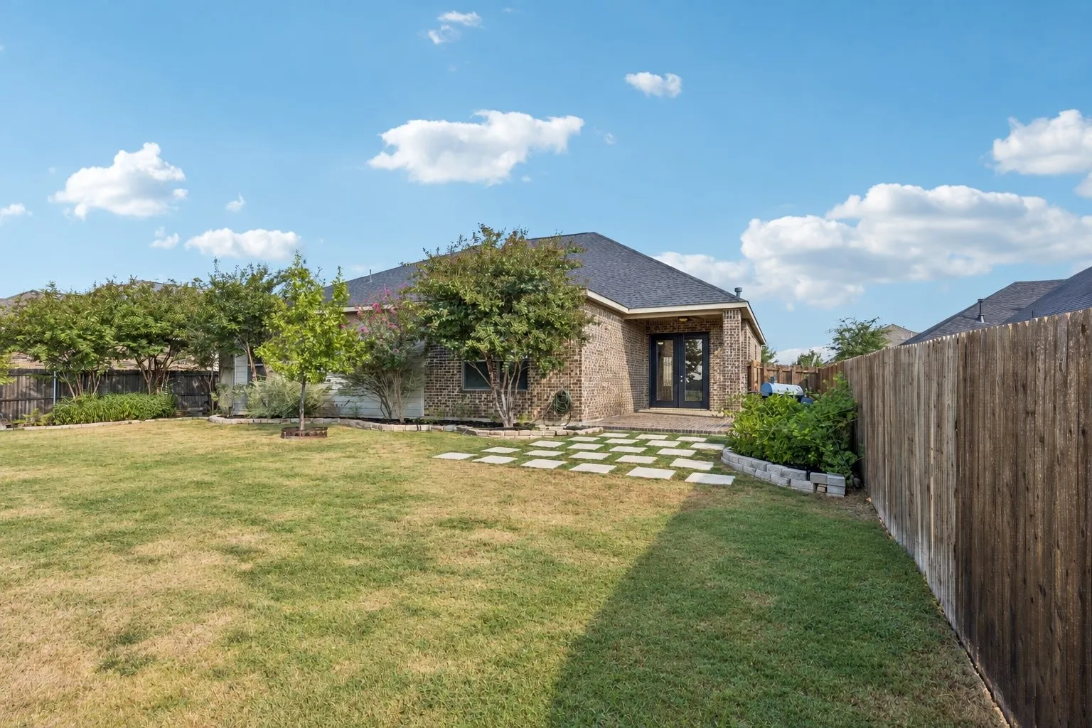 Expansive grassy backyard featuring a full privacy fence, brick exterior, and a stone paver patio