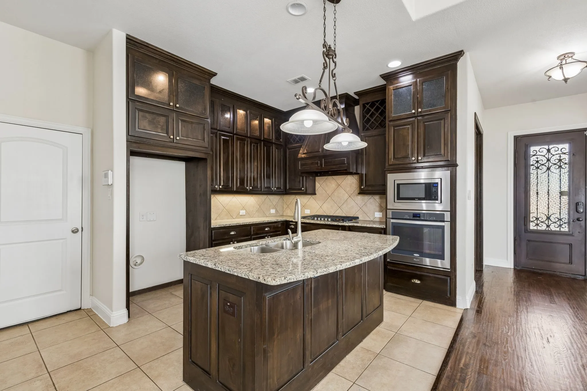 Kitchen featuring dark wood cabinetry, a central island with a granite countertop and sink, stainless steel appliances including a built-in oven and microwave, and a tiled backsplash