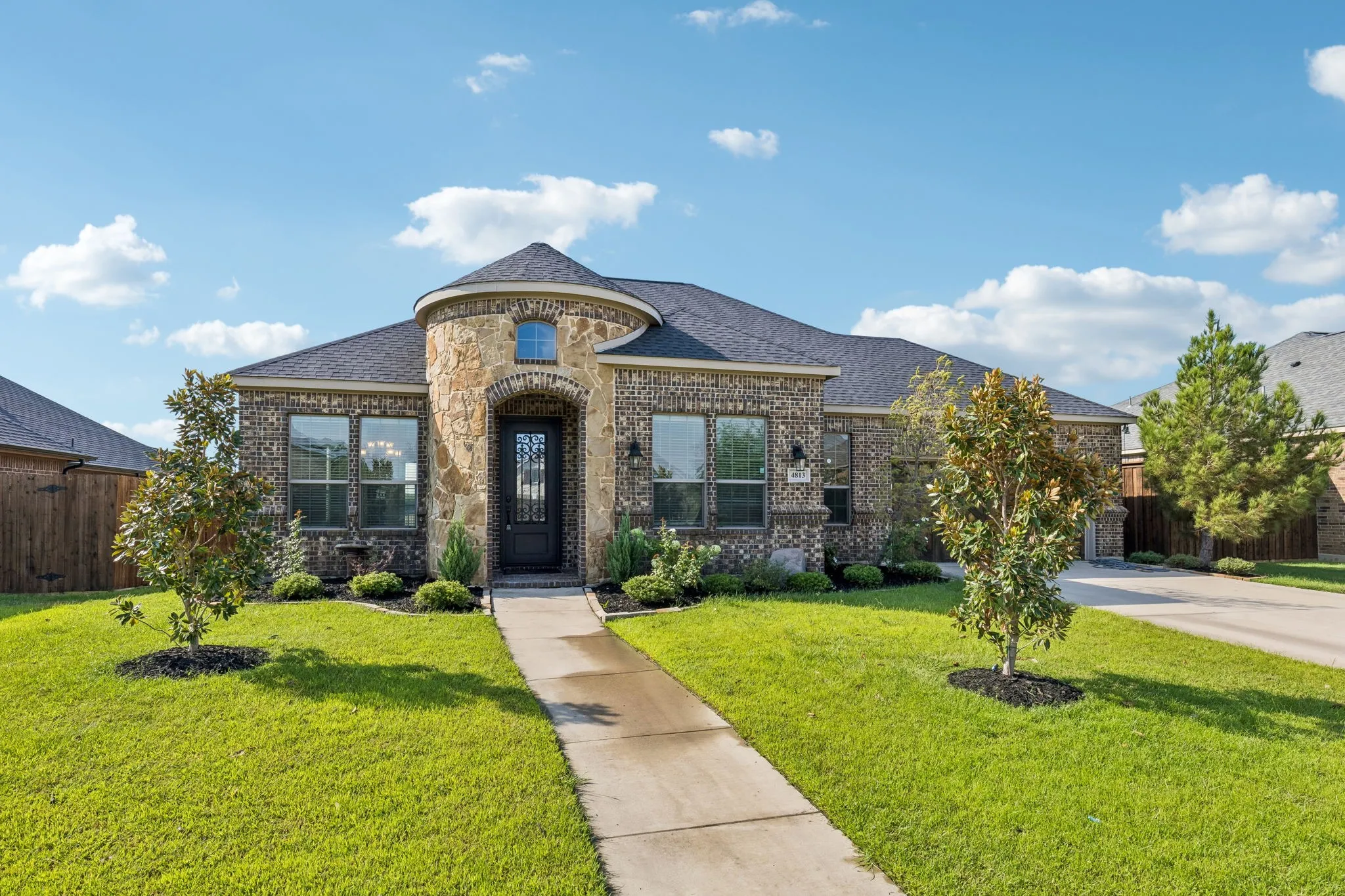 Stone and brick exterior with a turret-style entry, a dark shingle roof, a concrete walkway, and a landscaped front yard