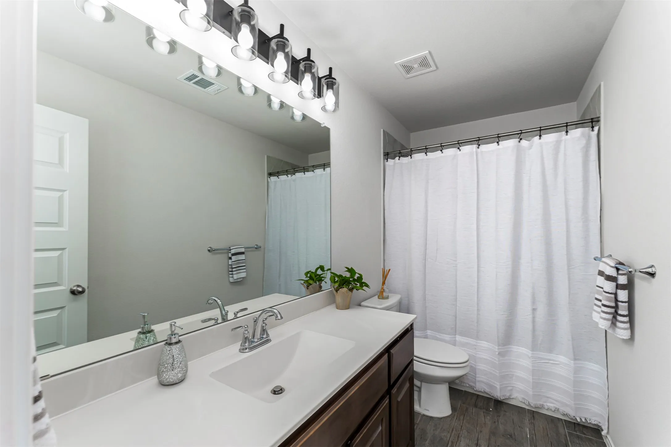 Bathroom featuring a single vanity with a white countertop, dark wood-finish cabinetry, a large wall-mounted mirror, a modern light fixture, and wood-finish flooring