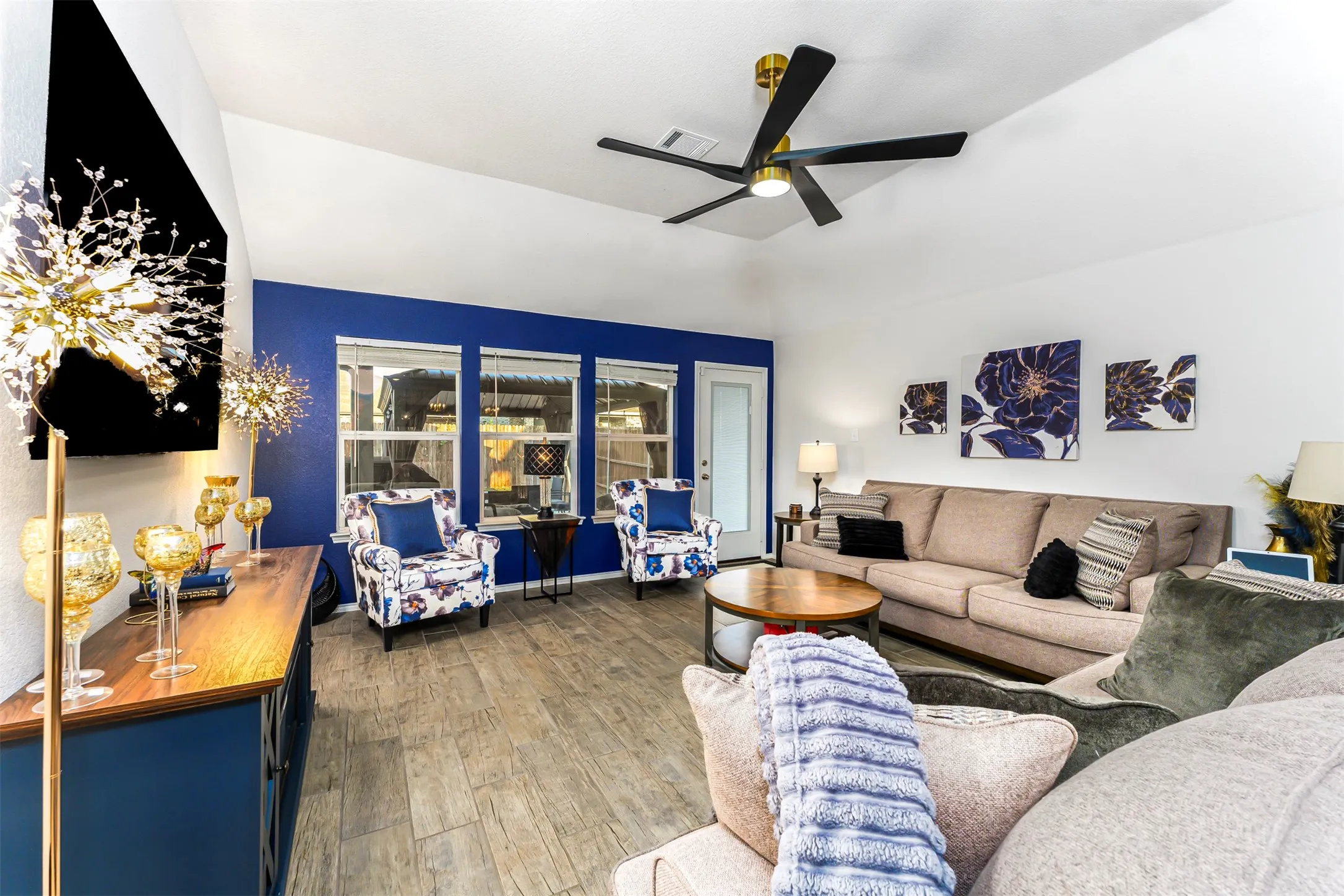 Vaulted ceiling with a black and gold ceiling fan, wood-finish flooring, accent wall, multiple windows, and a glass-paneled exterior door