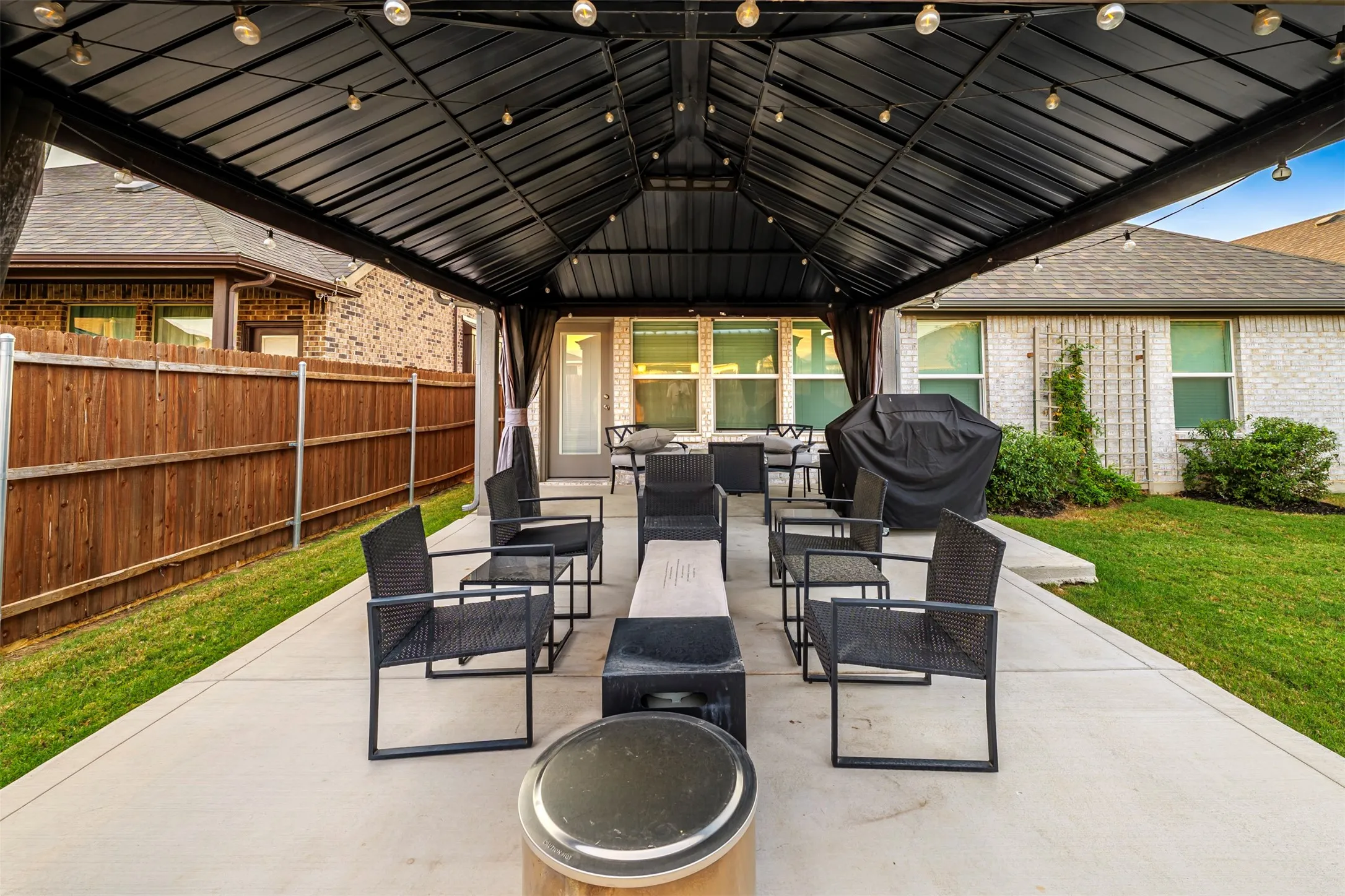 Covered patio structure with integrated string lighting, concrete patio, privacy fencing, and a surrounding lawn area