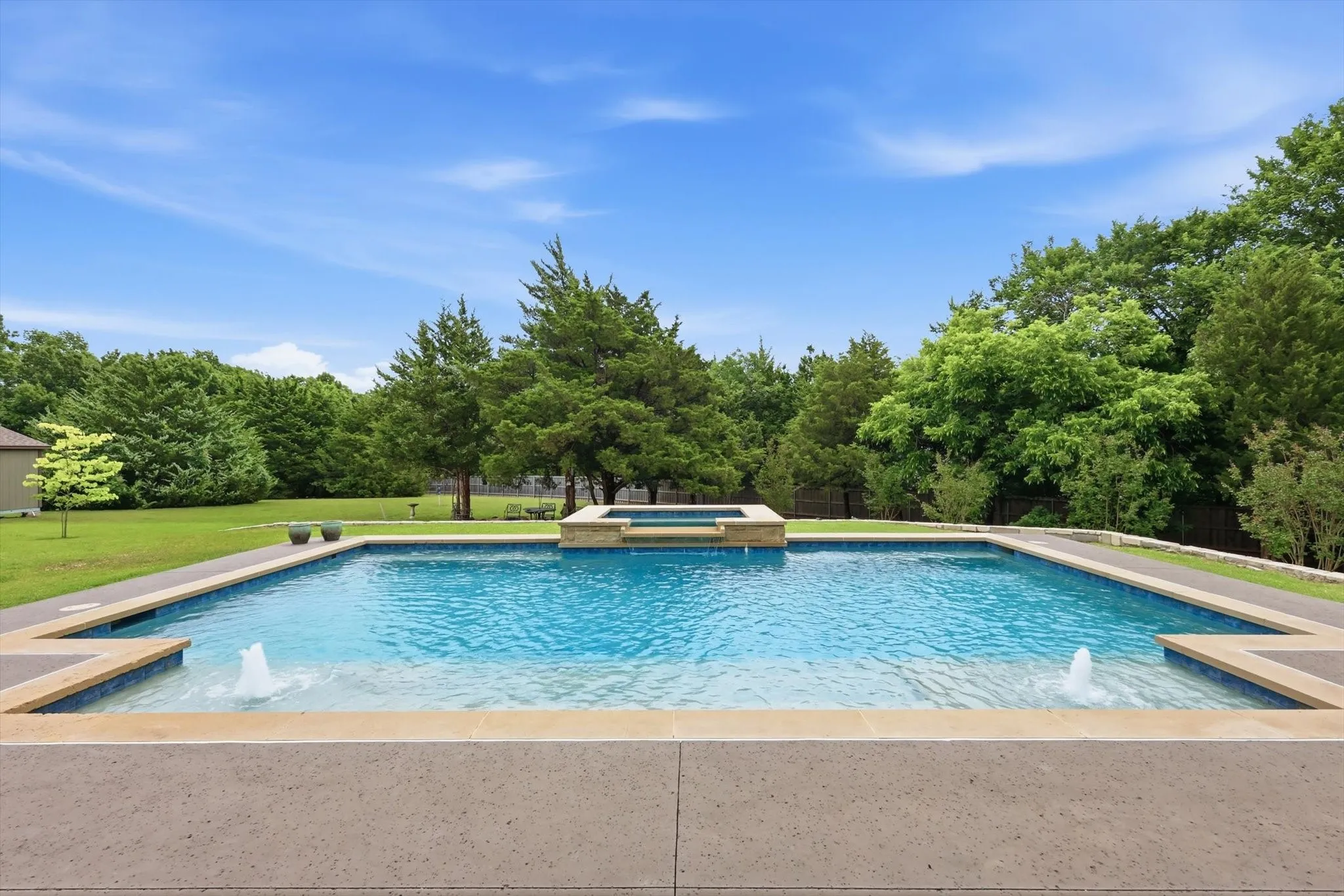 Expansive backyard featuring a large swimming pool, integrated spa, and dual water features, surrounded by a concrete deck and lush green lawn