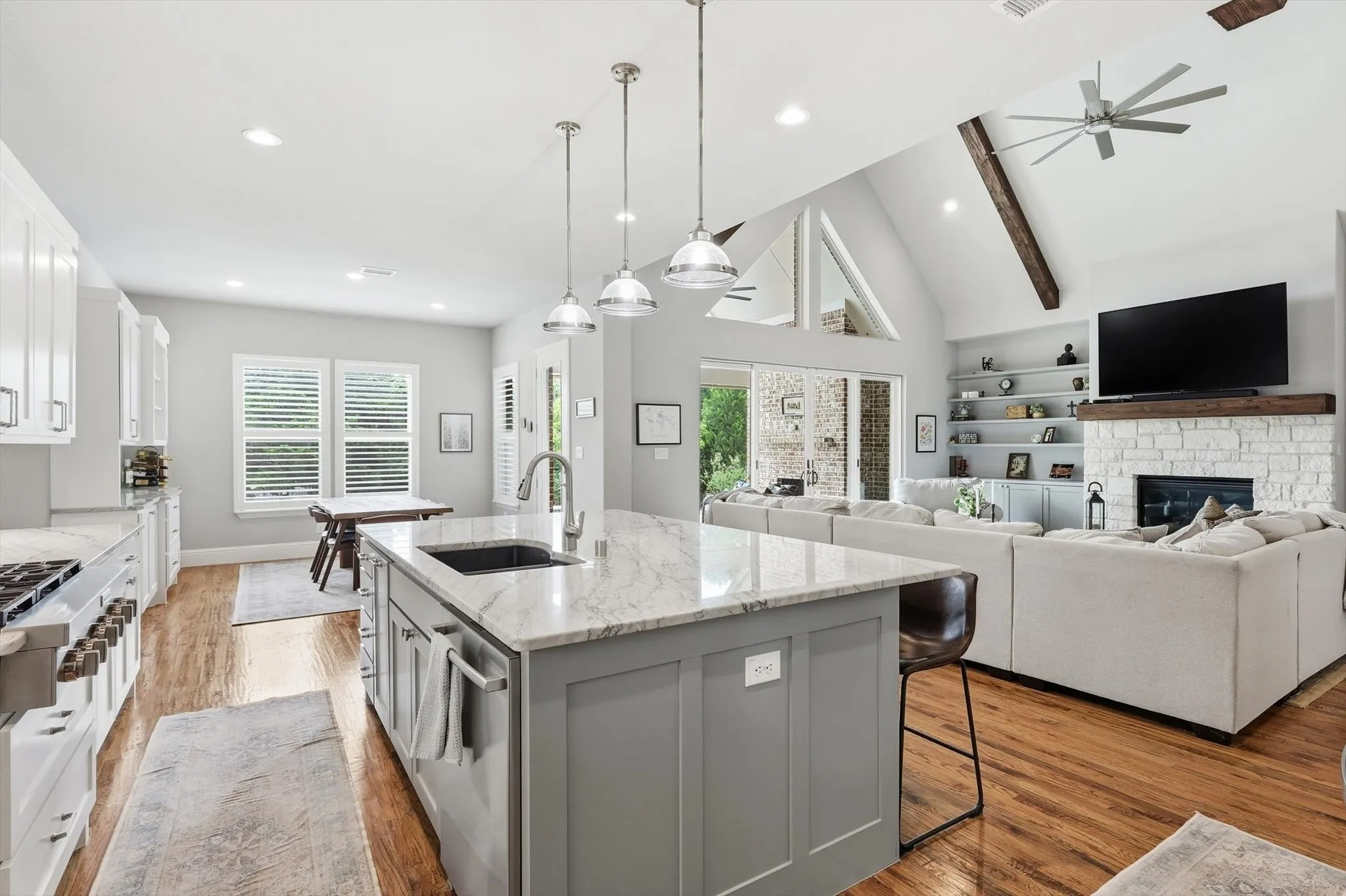 Open-concept living space featuring a vaulted ceiling with exposed beams, wood-finish flooring, a kitchen island with pendant lighting, a stone fireplace, and built-in shelving