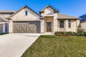 Brick exterior with stone accents, two-car garage with decorative crossbuck doors, concrete driveway, front entry door, and landscaped planting beds