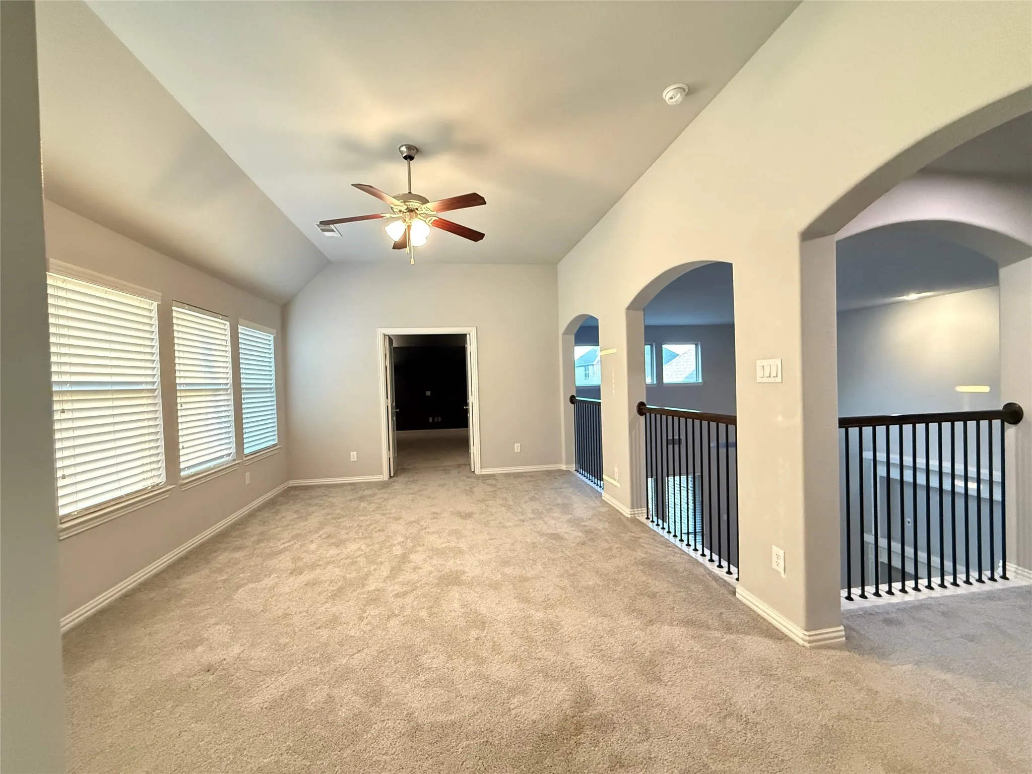 Carpeted room featuring a vaulted ceiling with a ceiling fan, multiple windows with blinds, a doorway, and arched openings to a balustrade with black spindles