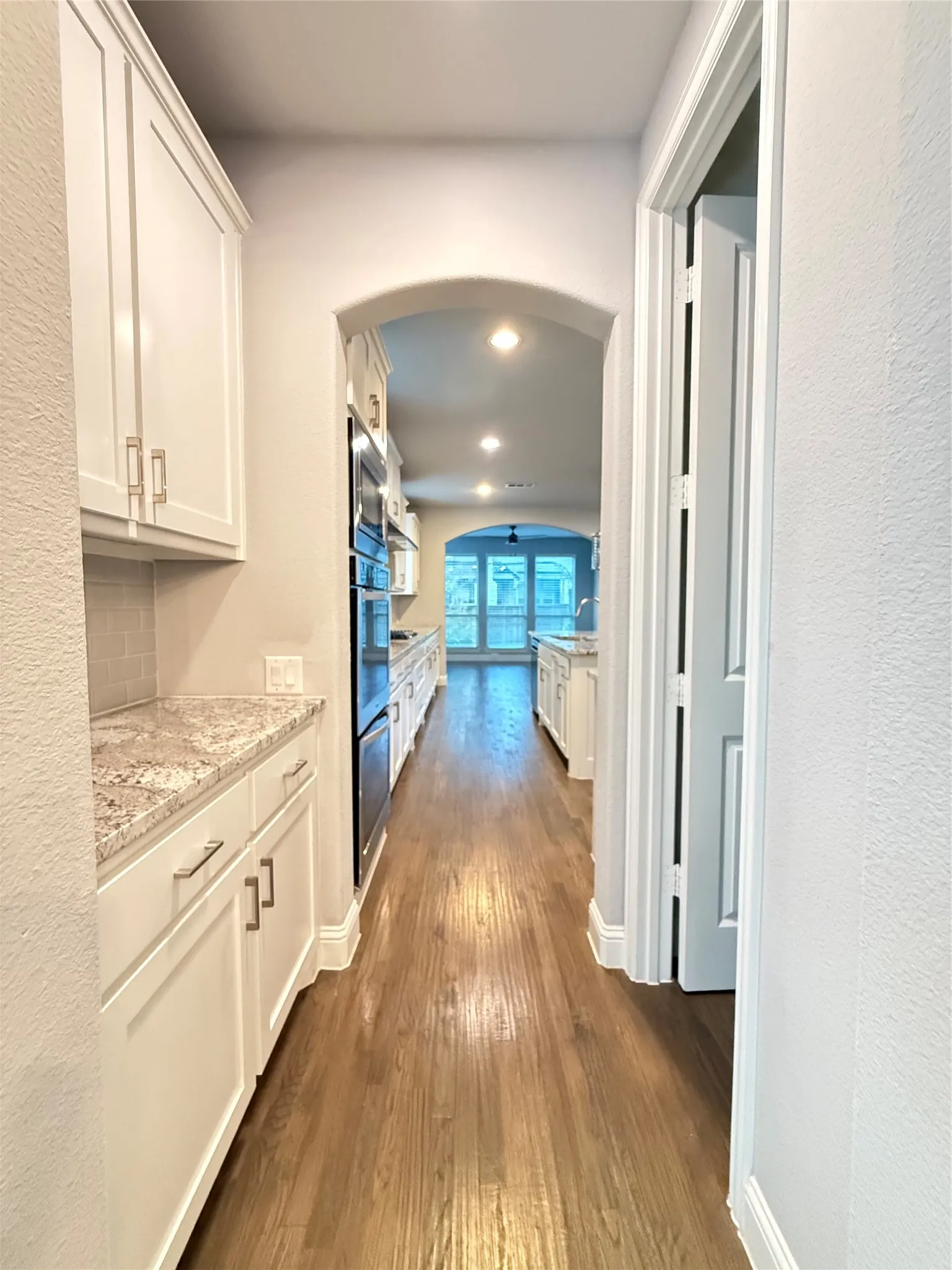 White shaker cabinetry with brushed nickel hardware, light-toned granite countertops, wood-finish flooring, built-in double ovens, and an arched entryway