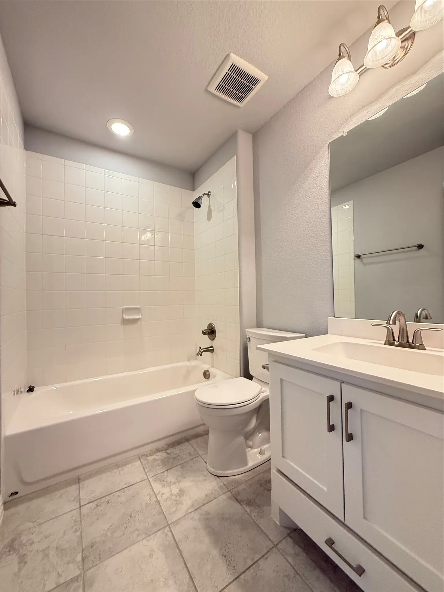 White subway tile surround, tile flooring, white vanity with shaker-style cabinetry, chrome faucet, and three-light vanity fixture