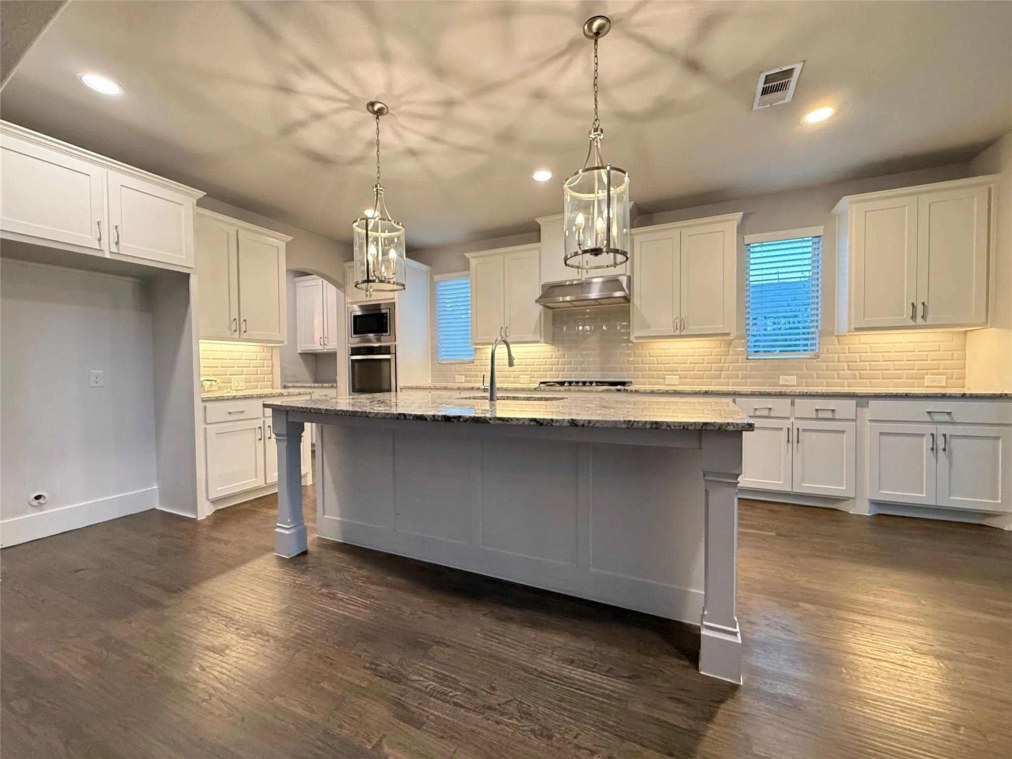 Central island with stone countertop, wood-finish flooring, white cabinetry, integrated stainless steel appliances, and pendant lighting fixtures