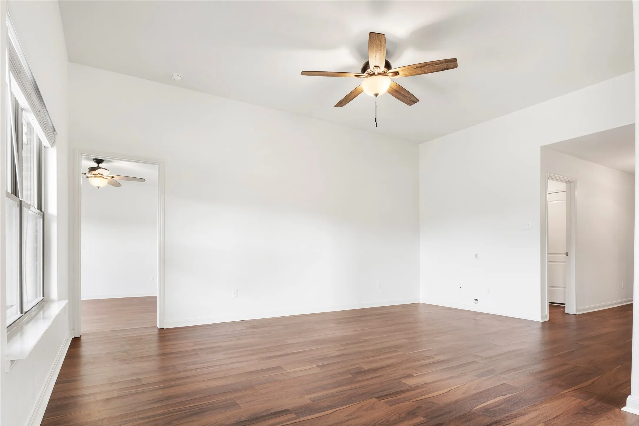 Vaulted ceiling, wood-finish LVP  flooring, ceiling fan with integrated lighting, multiple doorways, and a window with blinds. Viewed from the dining area.