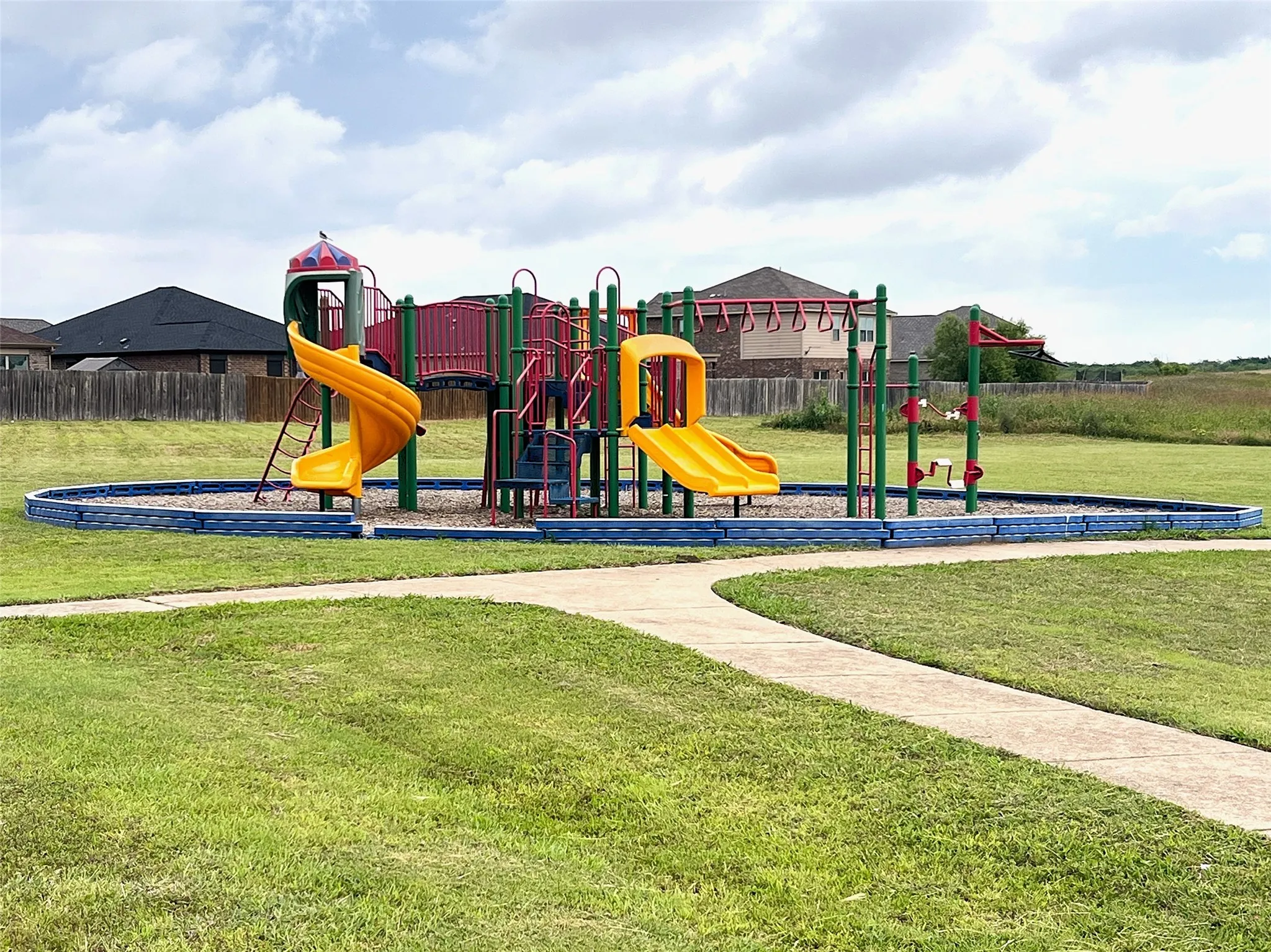 Recreational area featuring a multi-level play structure, two yellow slides, monkey bars, and a paved walking path