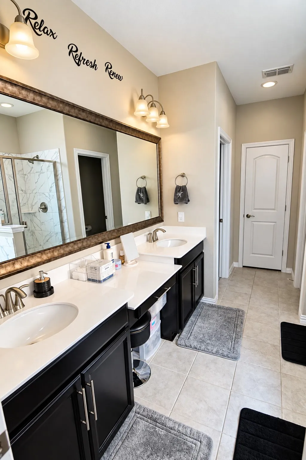 Primary bathroom, dual vanity with white undermount sinks and white countertops, dark wood-finish cabinetry with silver hardware, large framed mirror, and tiled flooring