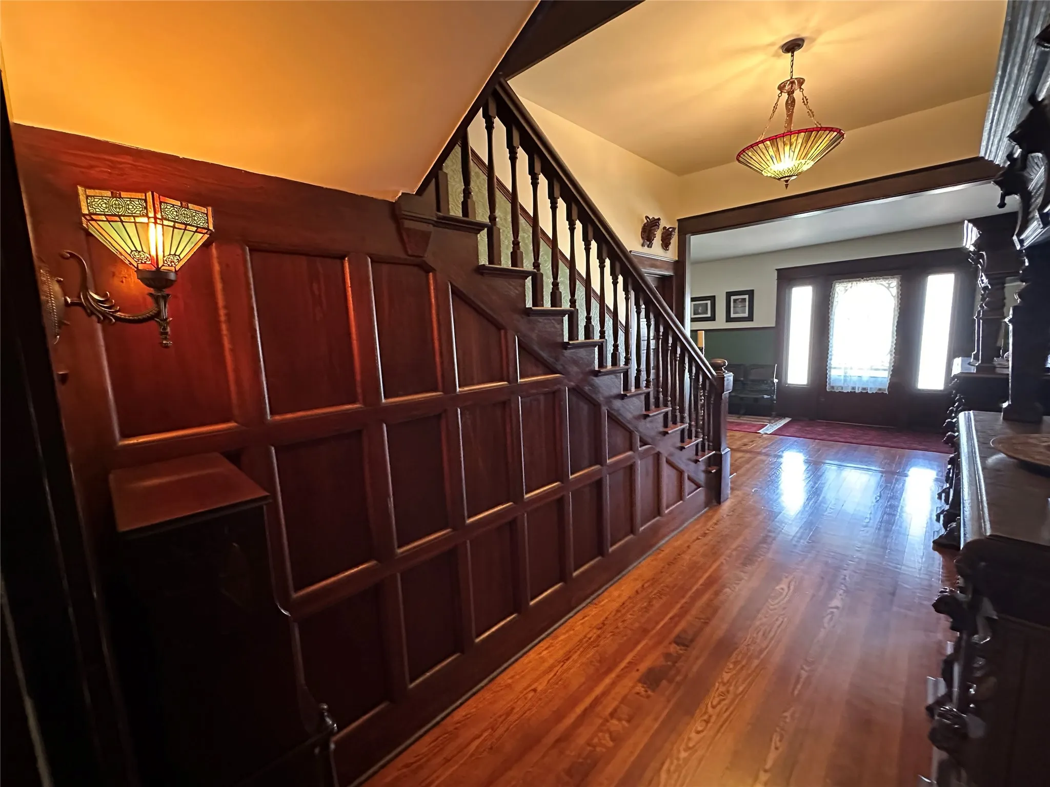 Hardwood flooring, wood paneling, wood staircase with balusters, stained-glass wall sconce, and a stained-glass chandelier