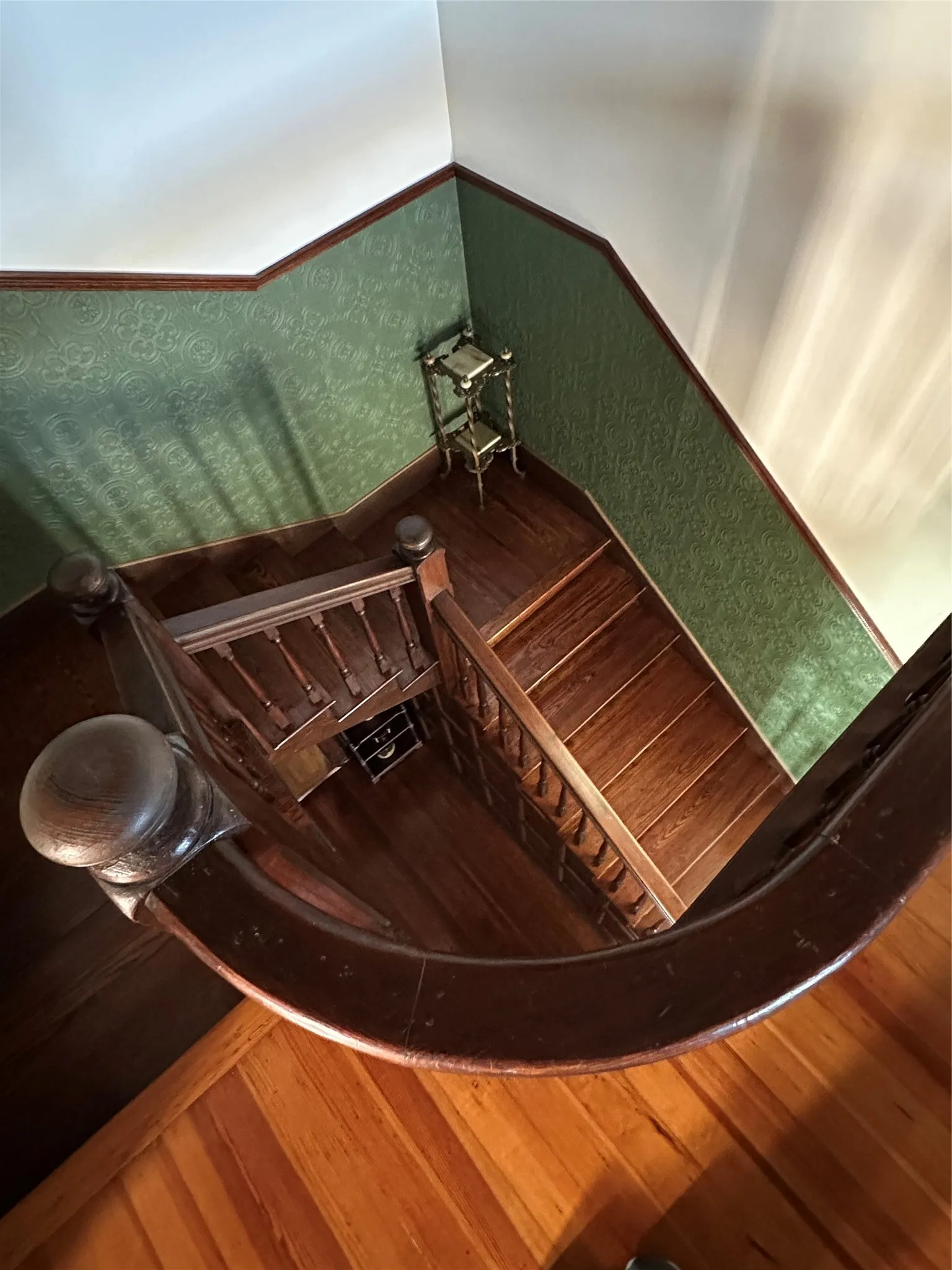 Hardwood staircase featuring a curved handrail, ornate balusters, wood-finish flooring, and decorative green wallpaper