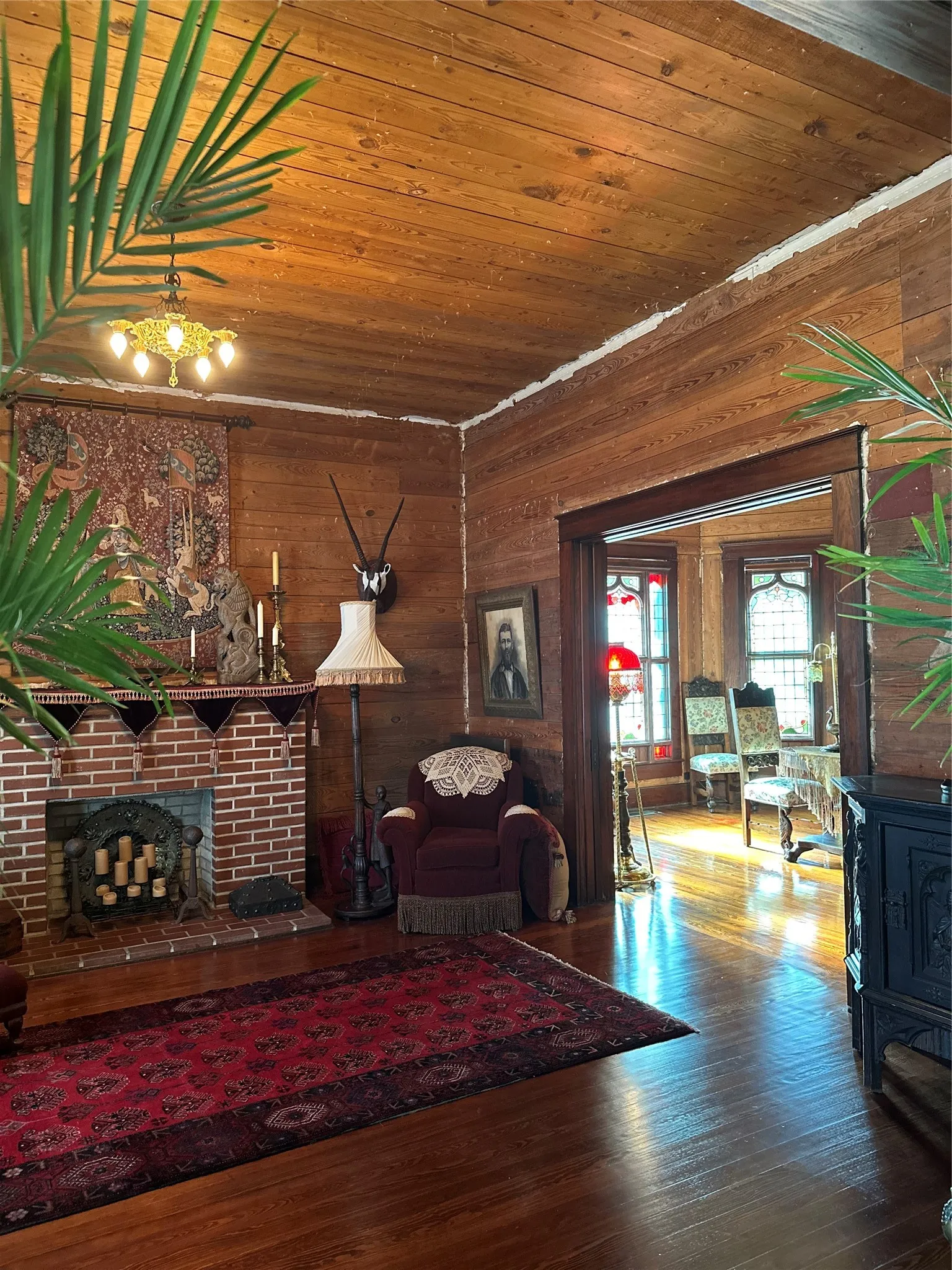 Wood-paneled walls and ceiling, brick fireplace with mantel, polished wood-finish flooring, and an open doorway to an adjoining room