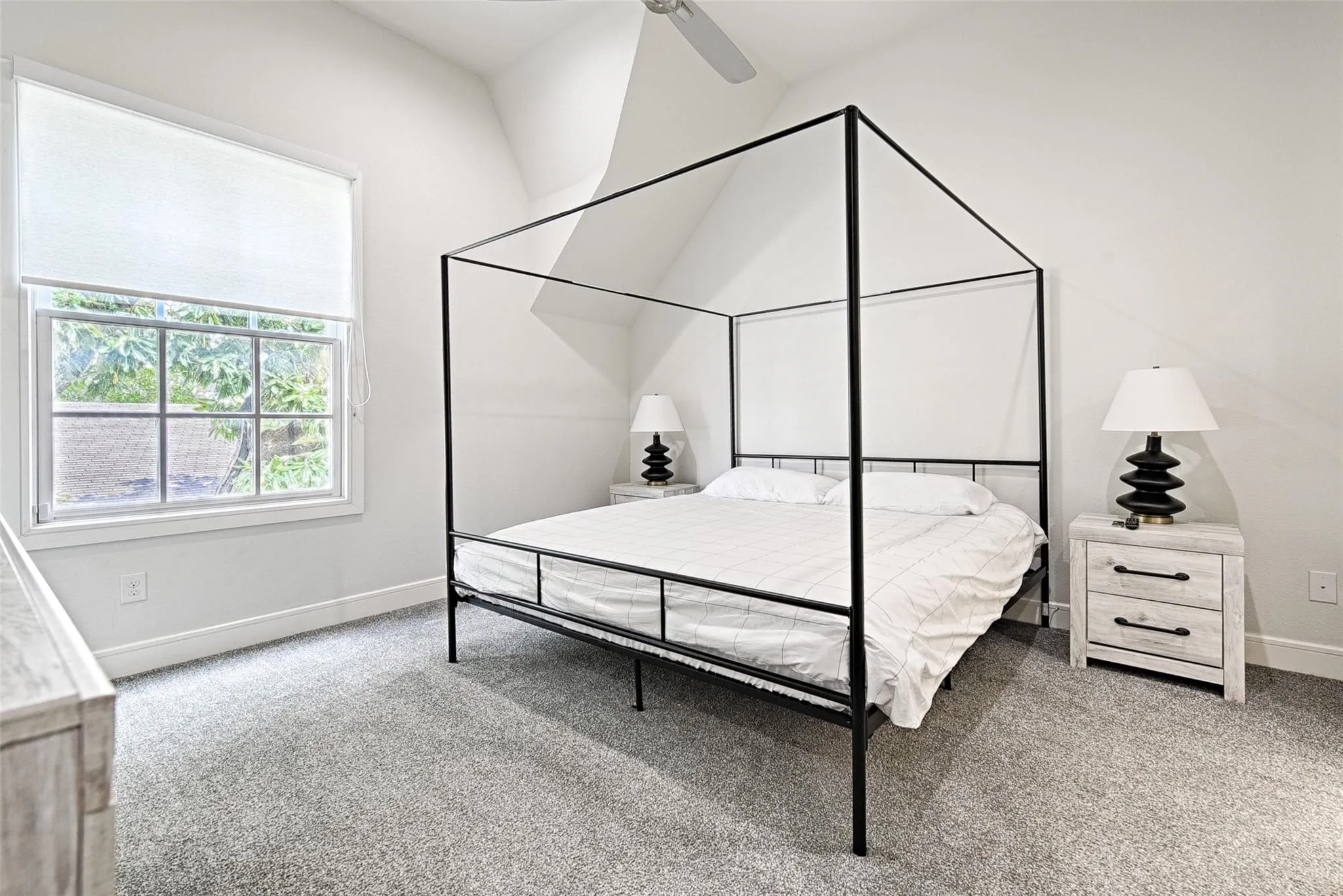 Vaulted ceiling with a ceiling fan, window with roller shade, and plush gray carpeting