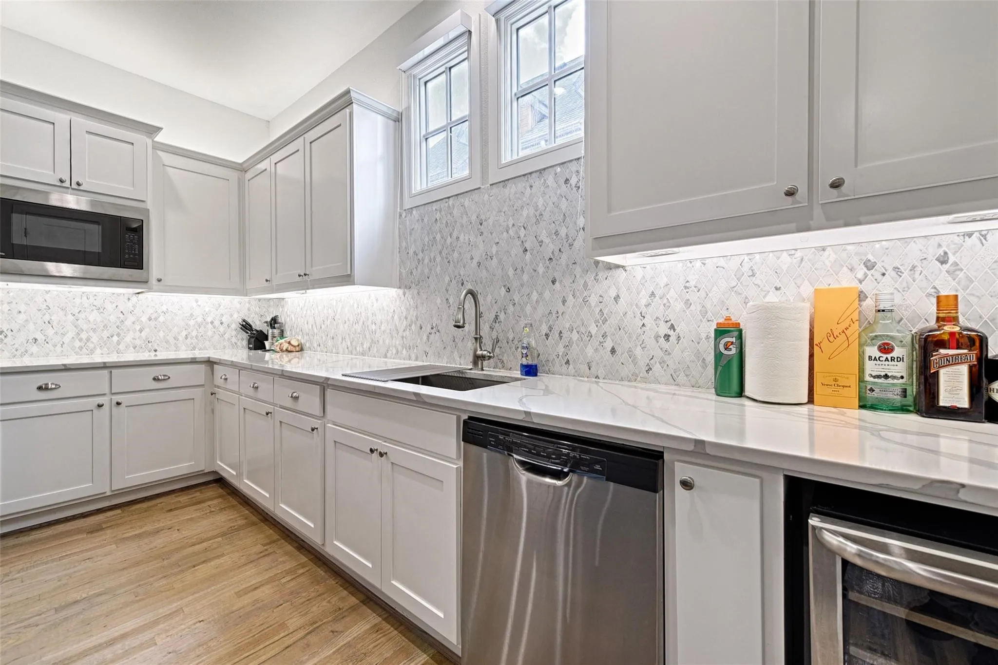 White shaker cabinetry, under-cabinet lighting, stainless steel appliances, patterned tile backsplash, and light-toned wood-finish flooring