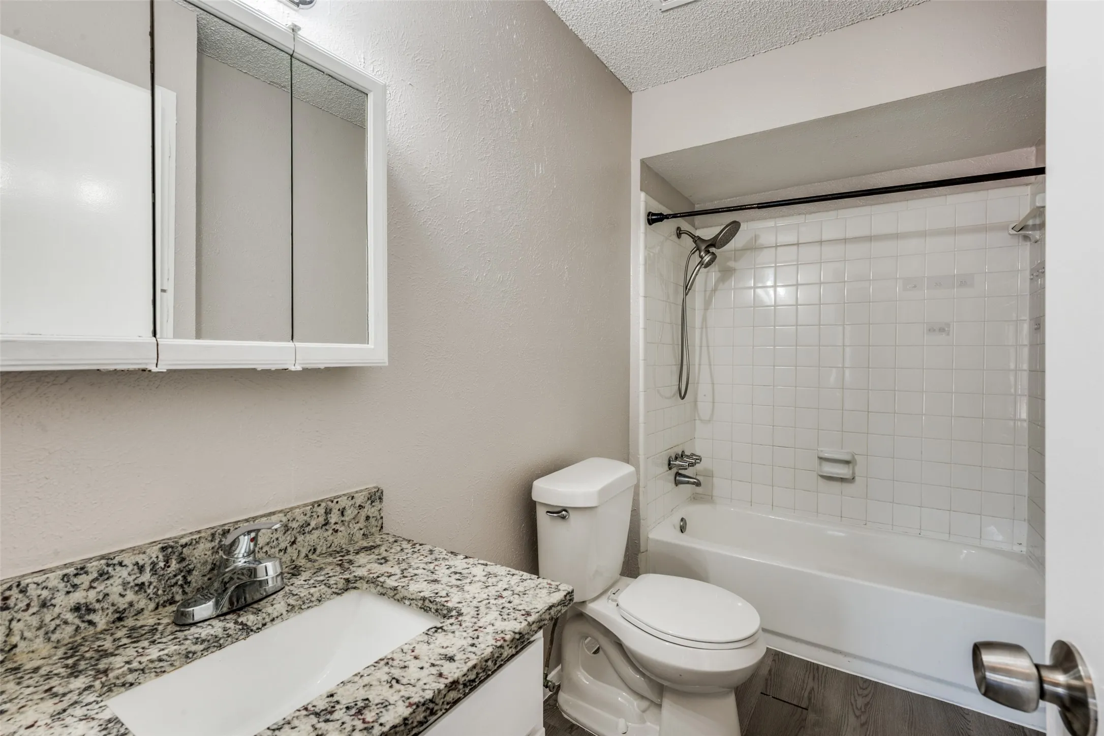 Bathroom featuring a vanity with a granular countertop, an undermount sink, a mirrored medicine cabinet, a toilet, and a tiled tub with a shower