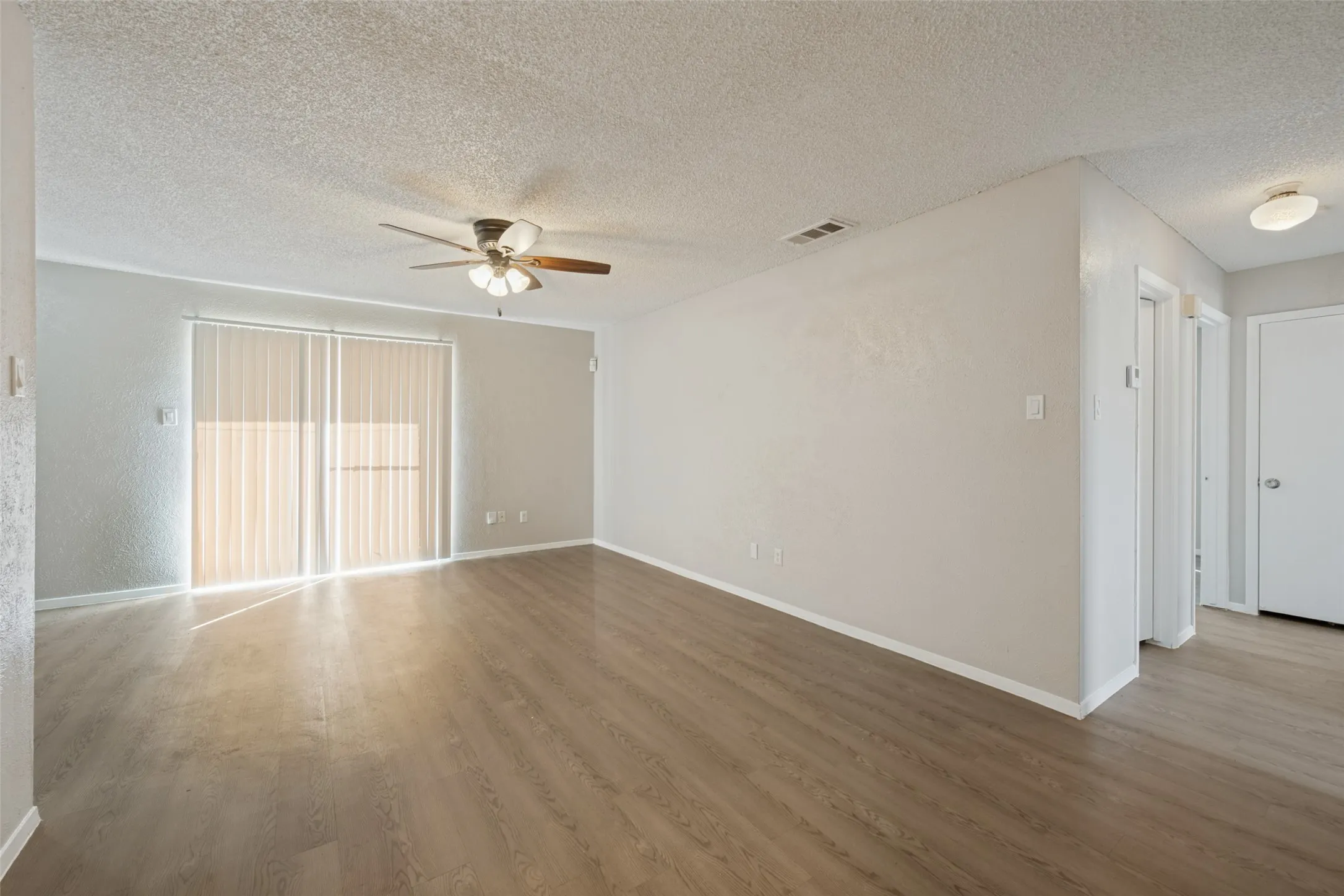 Spacious living area featuring wood-finish flooring, sliding glass doors with vertical blinds, a ceiling fan, and neutral paint