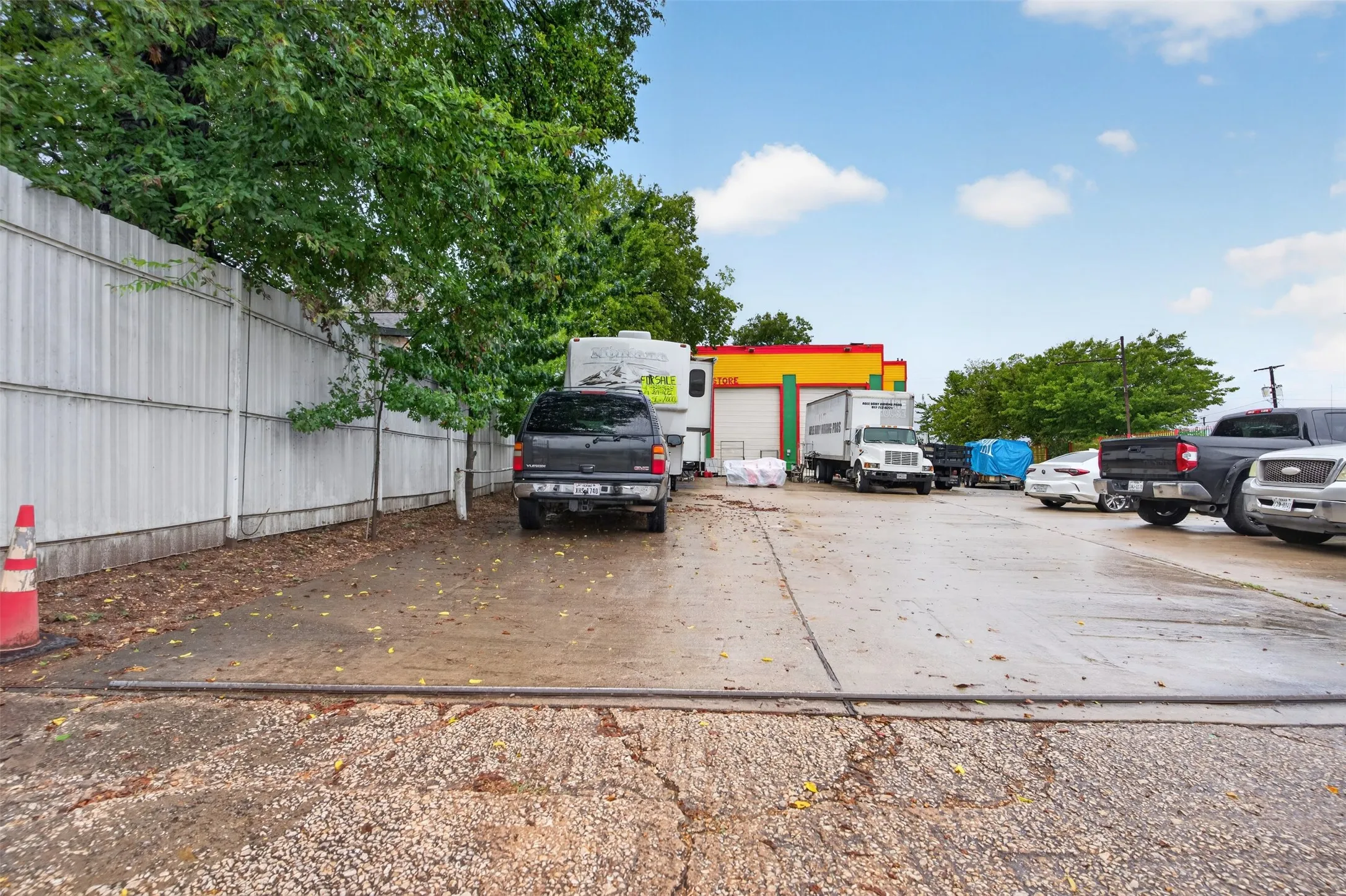 Paved parking area featuring a corrugated metal fence, mature trees, and a concrete surface