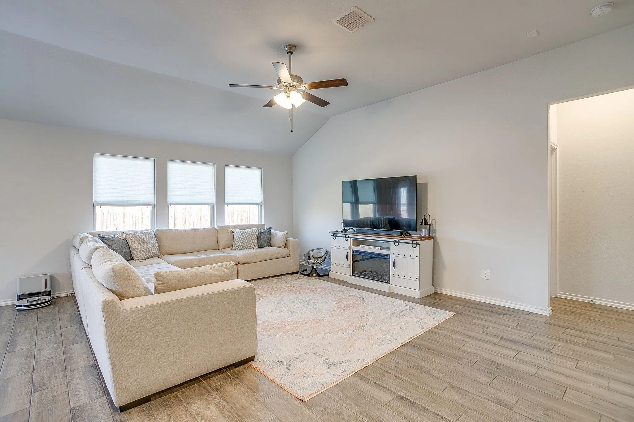 Vaulted ceiling with a ceiling fan, three windows with blinds, wood-finish flooring, and a built-in media console