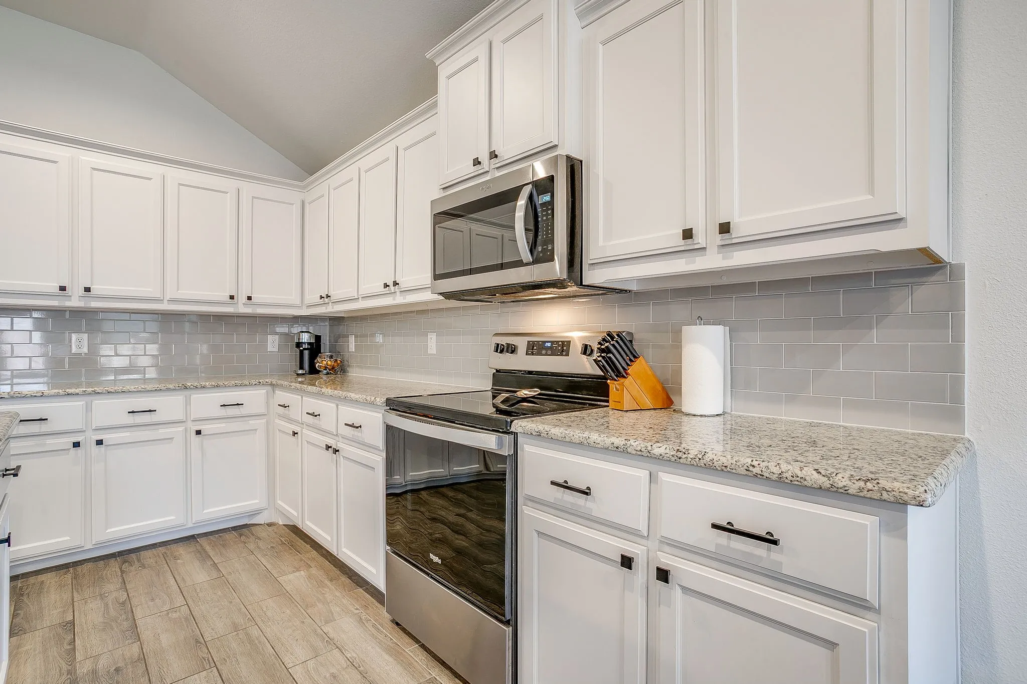 White cabinetry with dark hardware, stone countertops, stainless steel appliances, subway tile backsplash, and wood-finish flooring